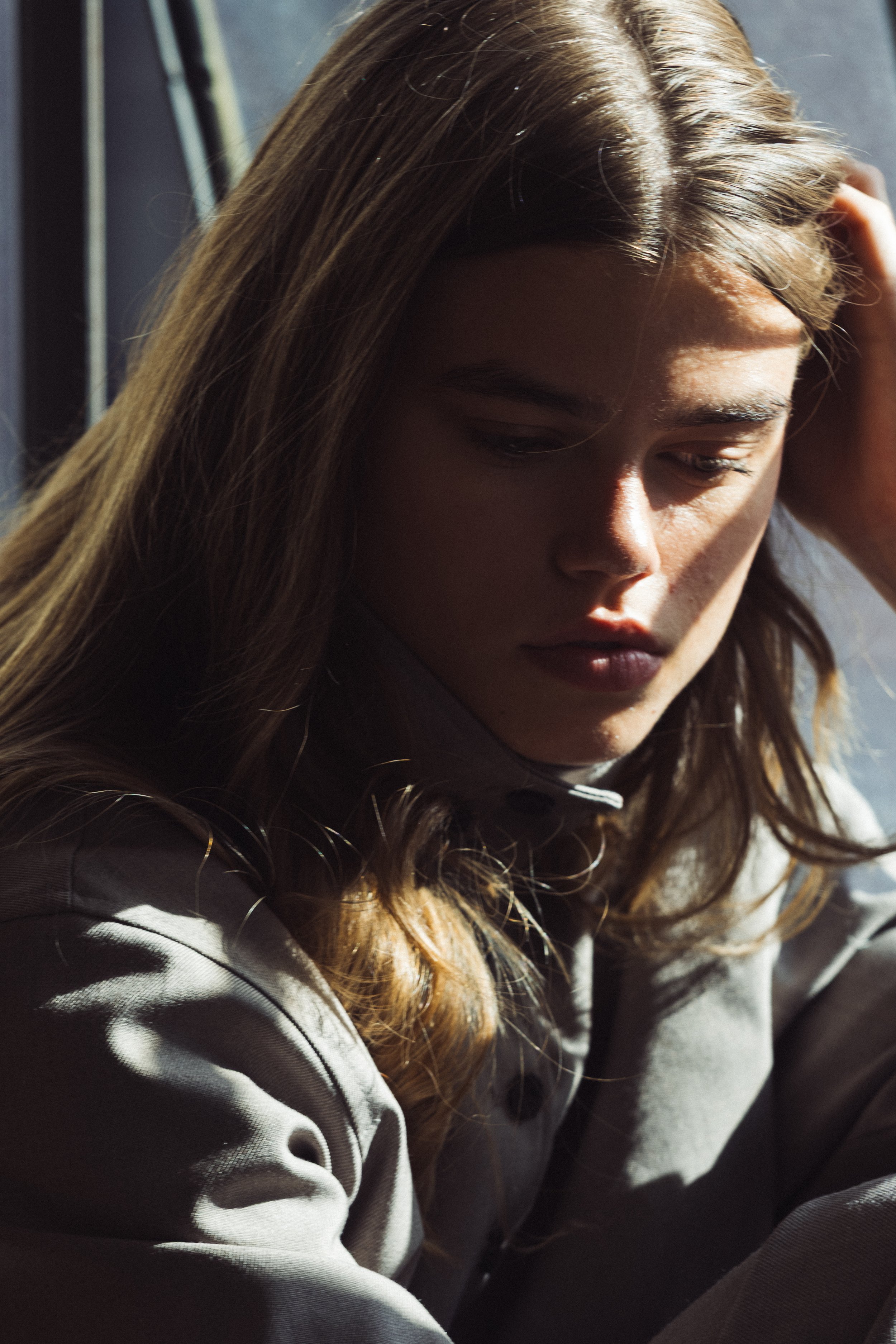 Close-up of a young woman with long light brown hair, looking down, sitting in natural sunlight.