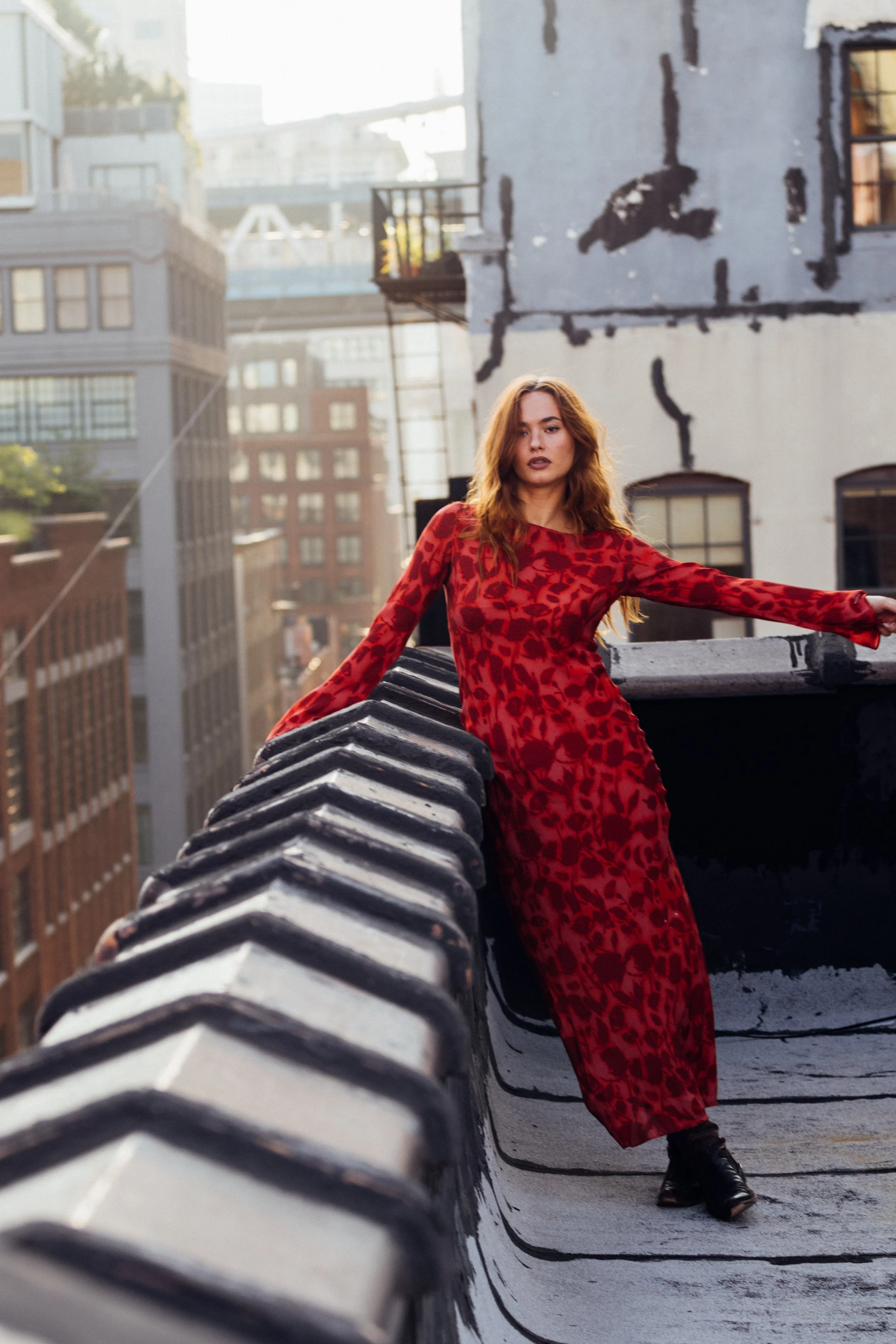 A woman with long red hair in a red patterned dress and black boots stands on a rooftop with city buildings in the background, leaning against a chimney with a serious expression.