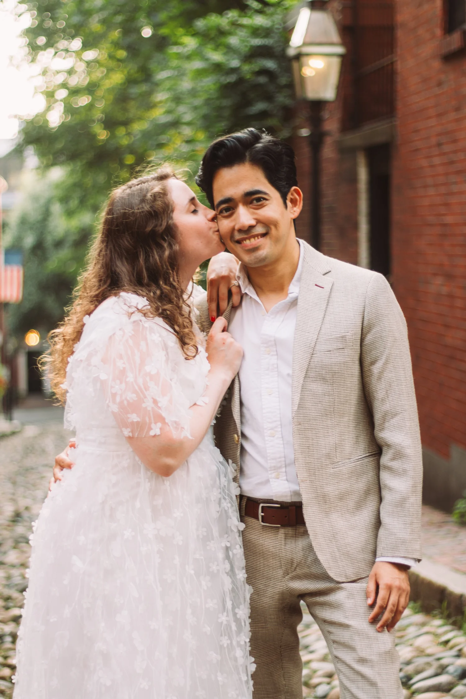 Maddy kissing Stefano on the cheek beside brick buildings and trees