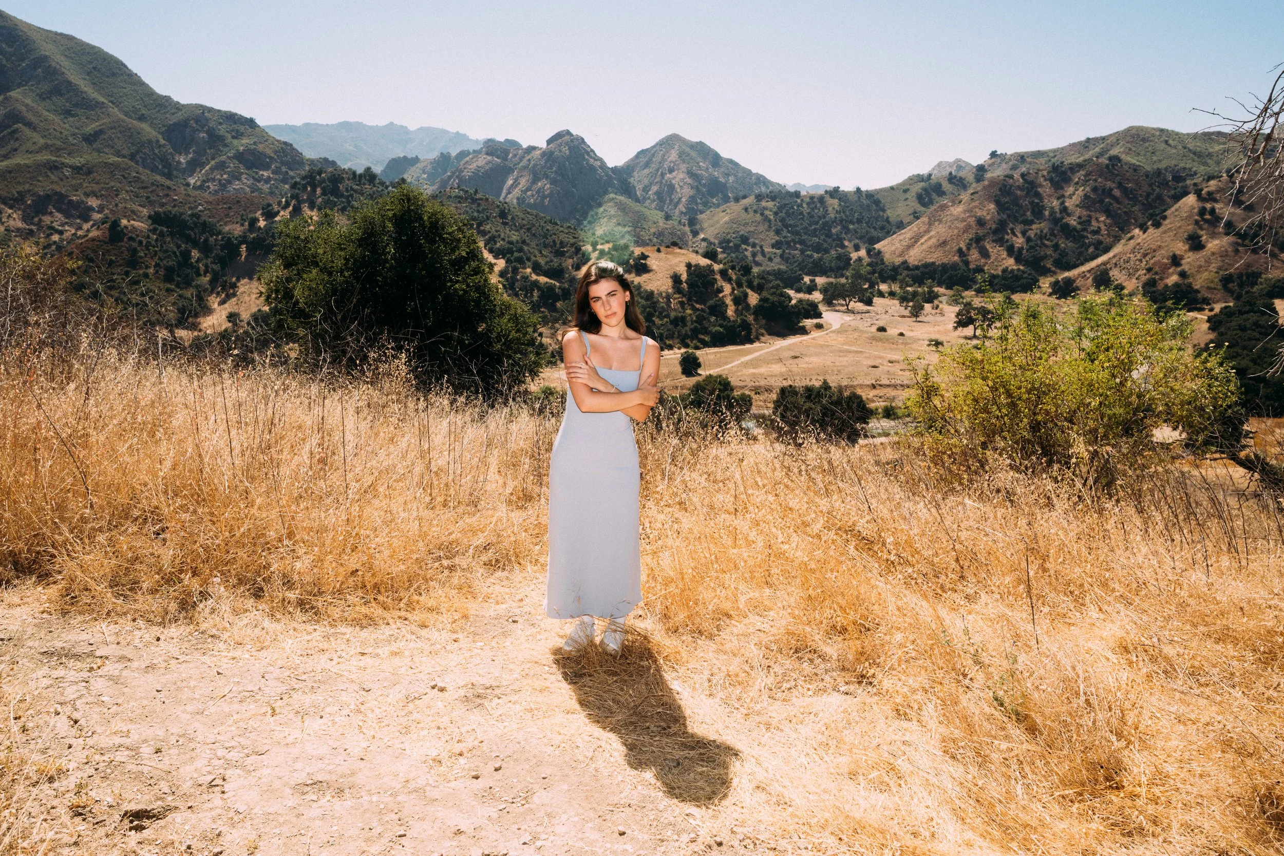 A young woman in a long, light-colored dress standing in a dry, grassy field with mountains in the background during daytime.