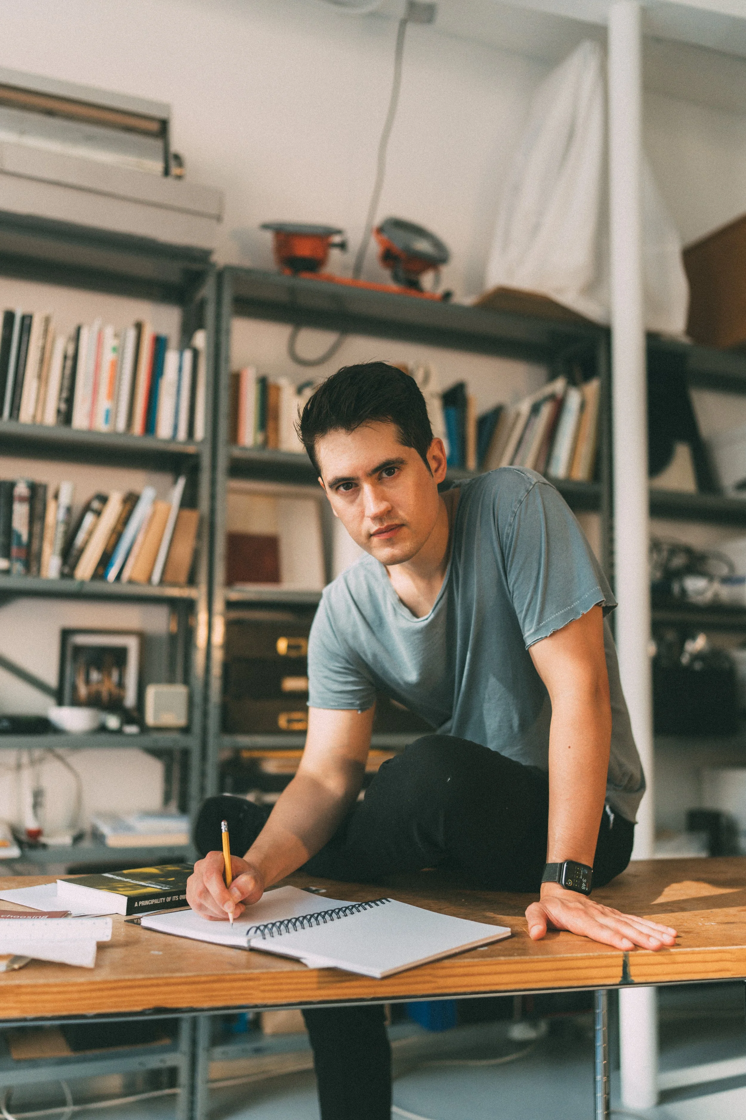 Young man with dark hair and wearing a gray t-shirt, sitting at a wooden table, writing in a notebook with a pencil, in a room with bookshelves filled with books and some personal items.
