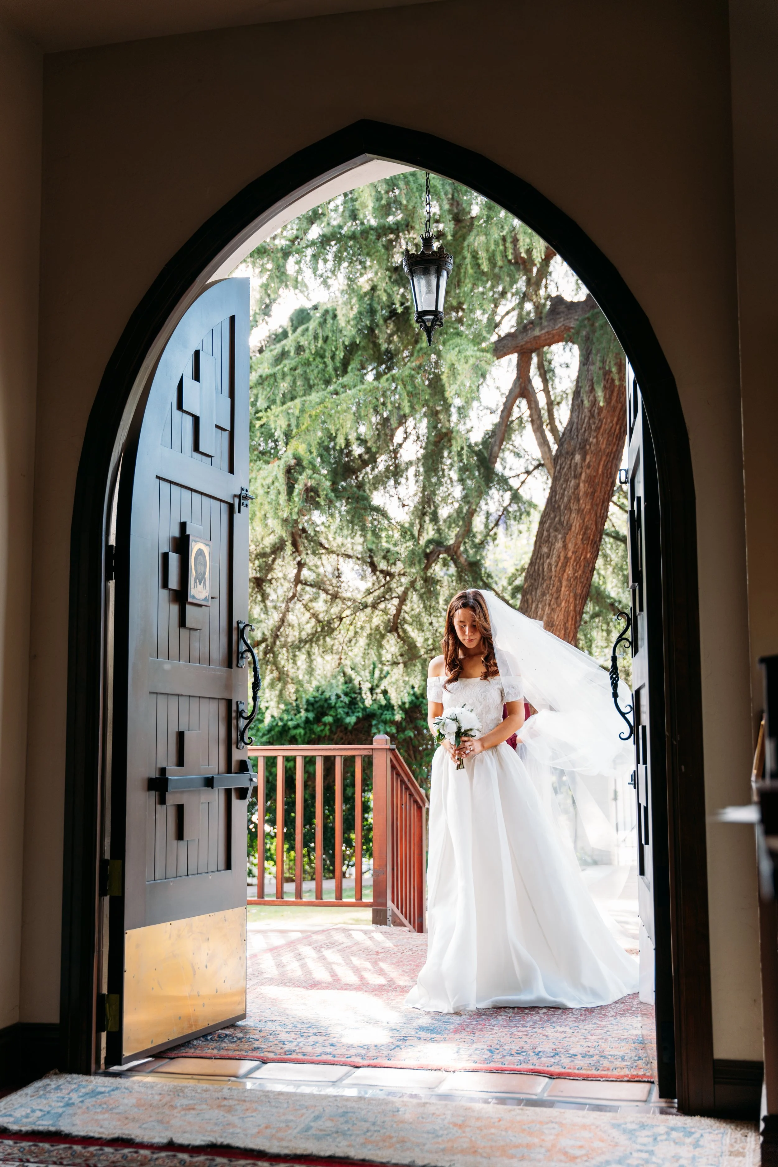 A bride in a white wedding dress holding a bouquet stands just outside a doorway, with a veil flowing in the breeze, framed by open dark wooden doors, with trees and a railing in the background.