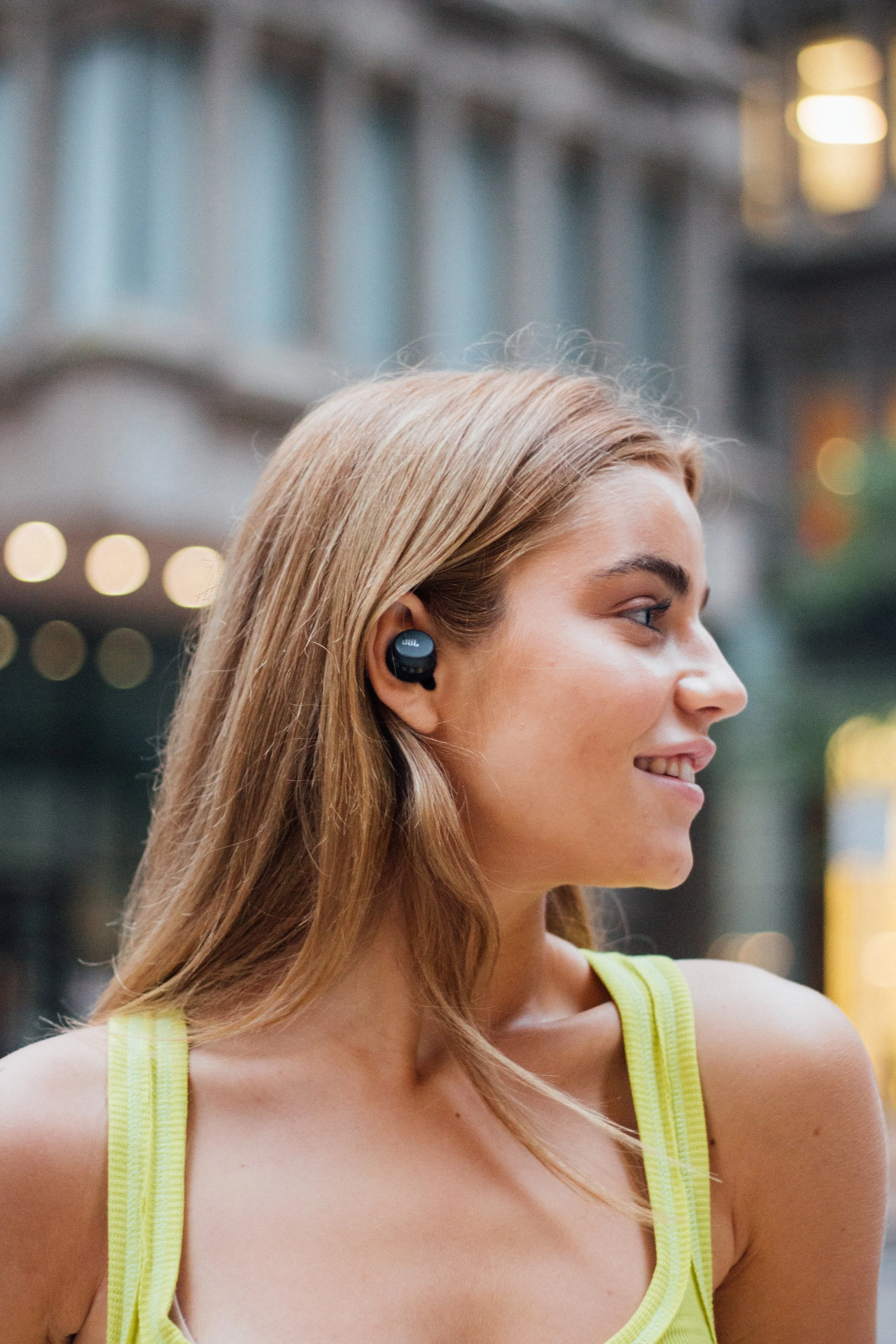 A young woman with blonde hair wearing wireless earbuds and a yellow tank top, smiling and looking to the side in an urban setting with blurred city lights in the background.