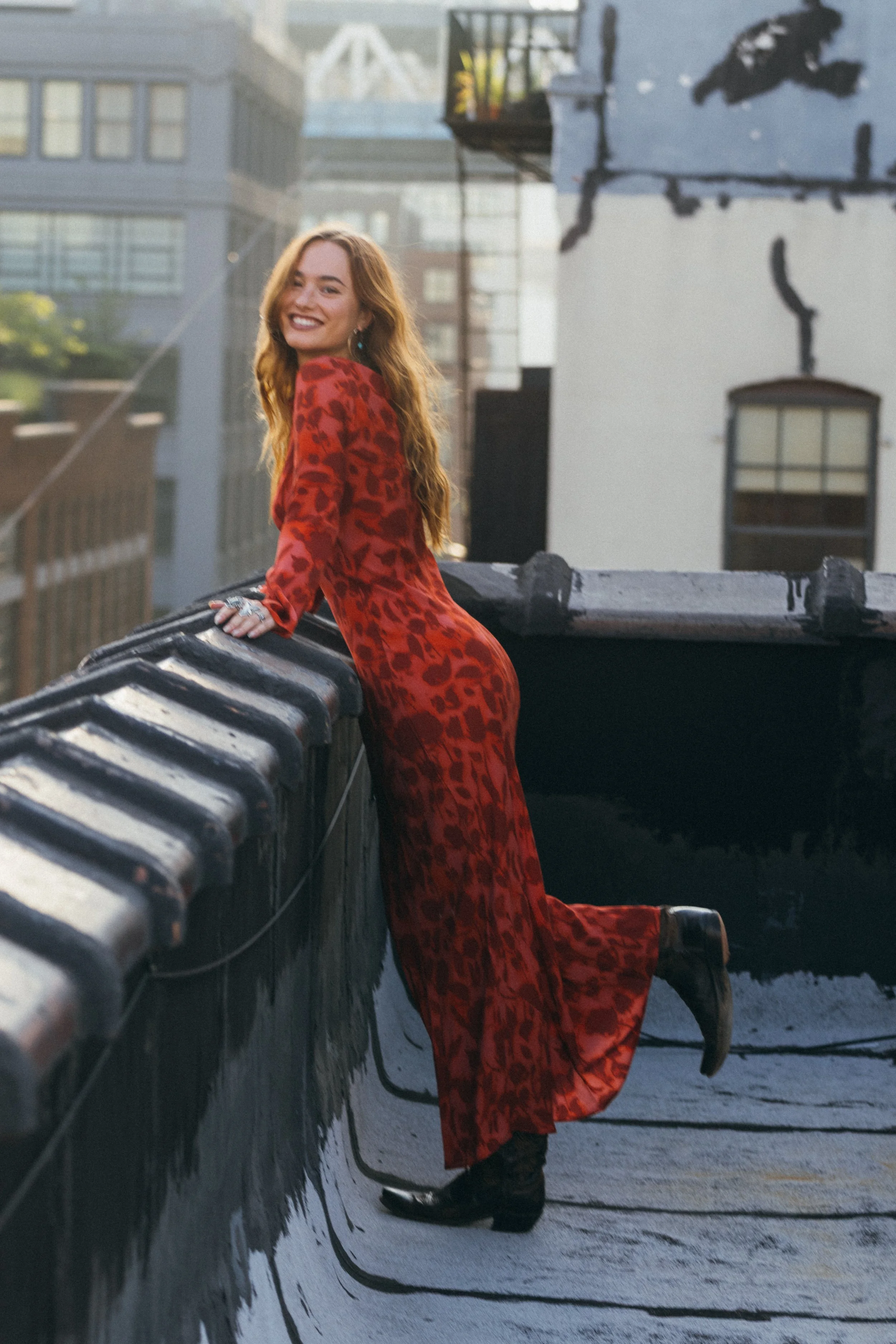Johnny in a red patterned dress leaning against a rooftop railing, with New York buildings behind her.