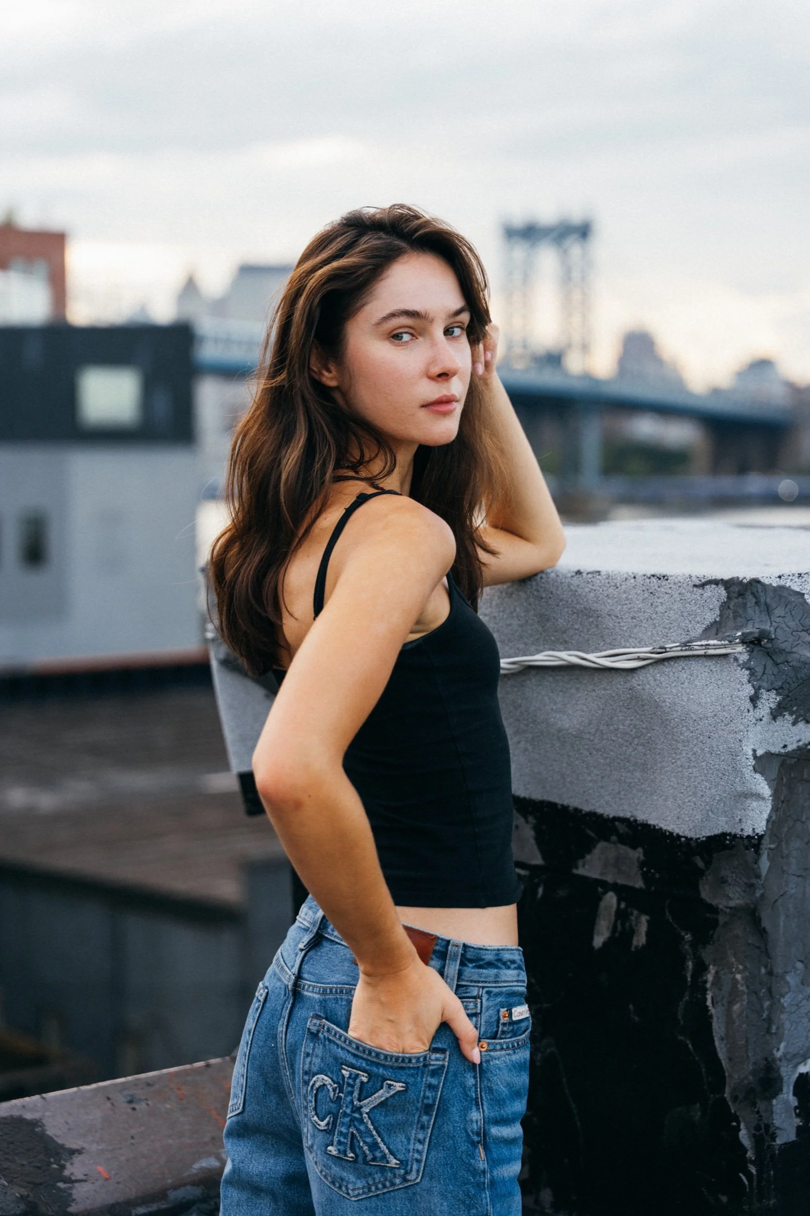 Model in a black tank and jeans looking back over one shoulder on a city rooftop