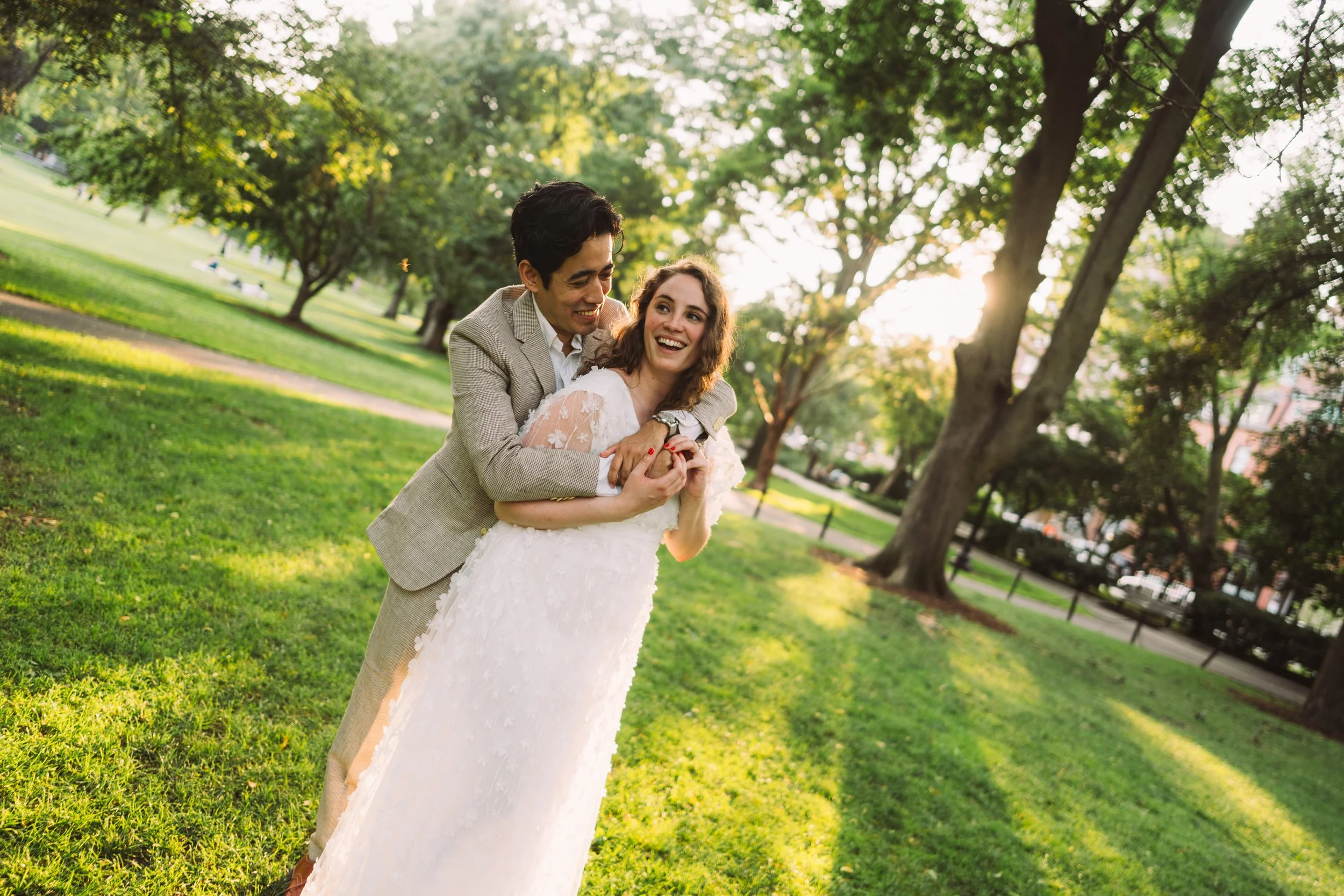 A man in a beige blazer and a woman in a white wedding dress smiling and embracing each other in a park with green grass and trees during sunset.