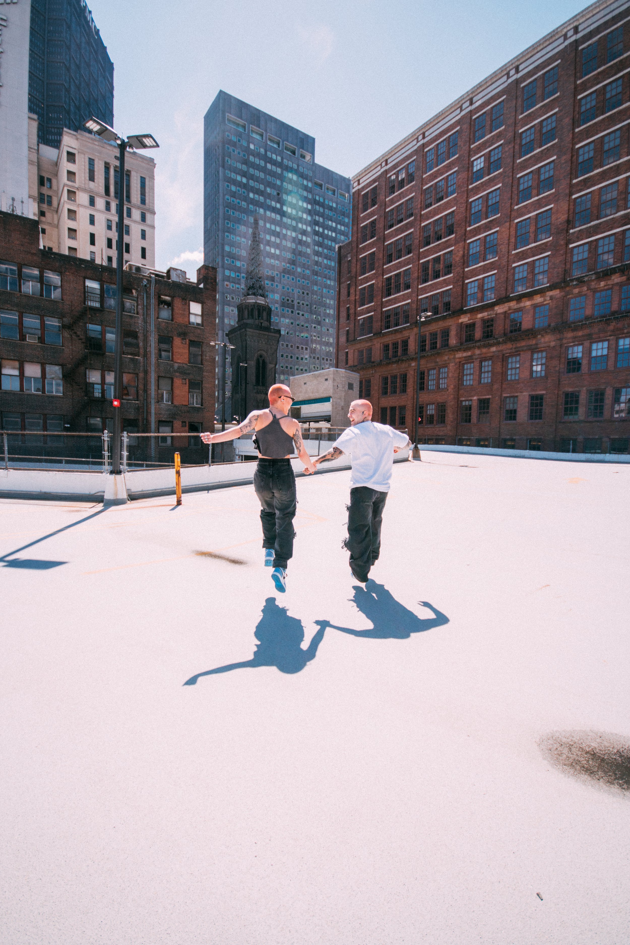 Two people holding hands and jumping in the air on a rooftop parking lot with New York City skyscrapers in the background.