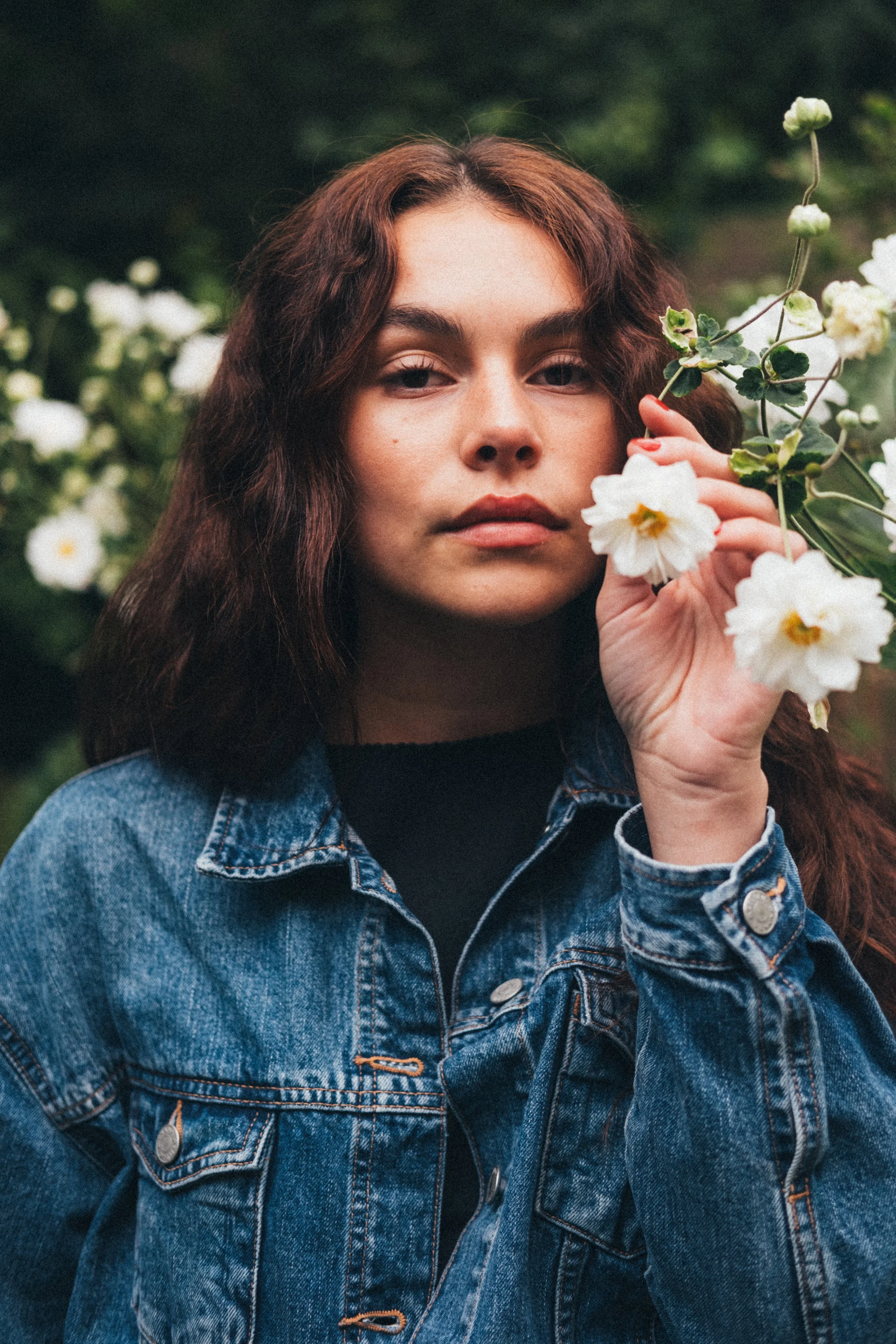 A young woman with long, curly, auburn hair, wearing a denim jacket, holding a white flower near her face, standing outdoors with a blurred background of greenery and white flowers.