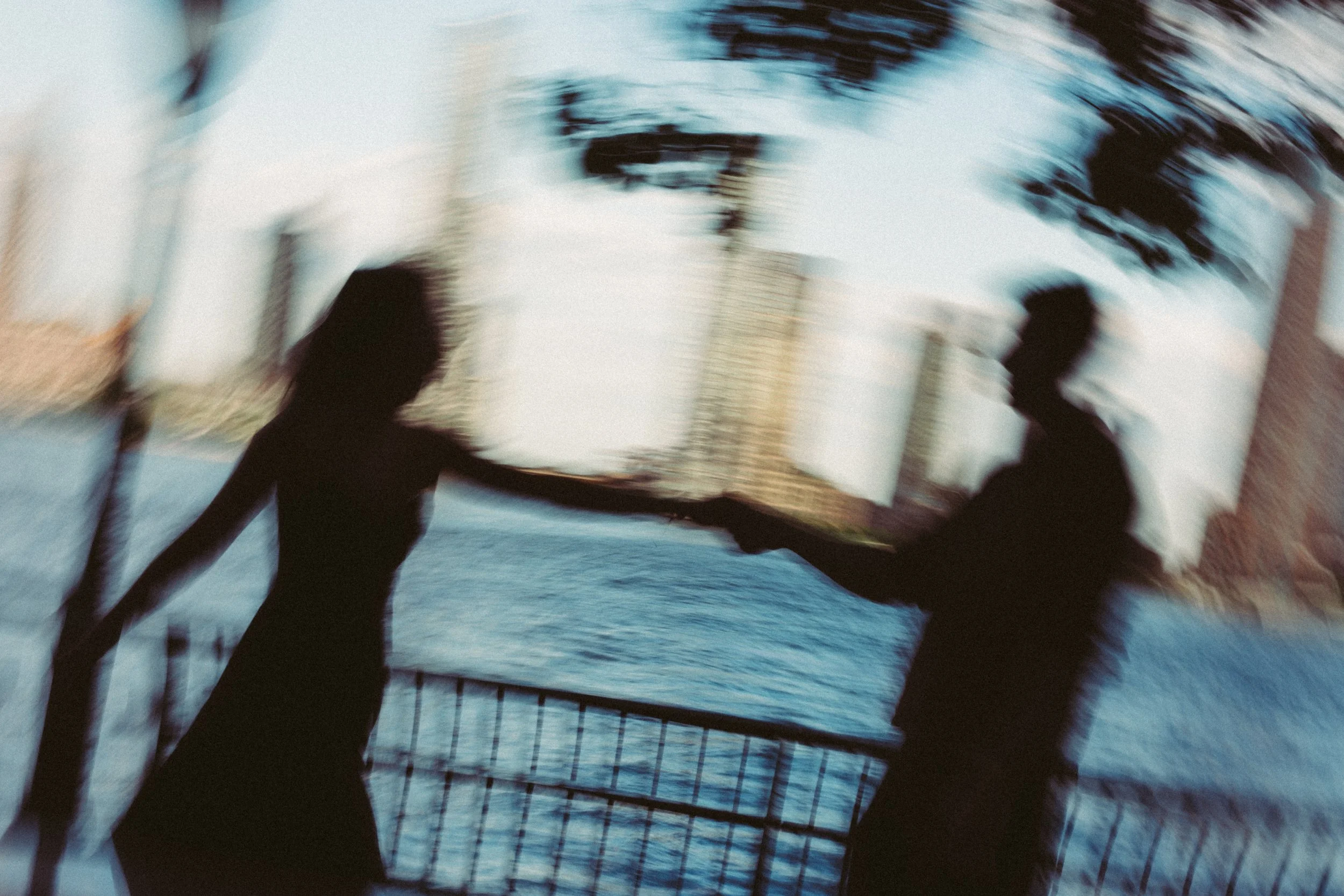 Two people holding hands while walking on a bridge or walkway over water, with trees and buildings in the background, blurred motion shot.