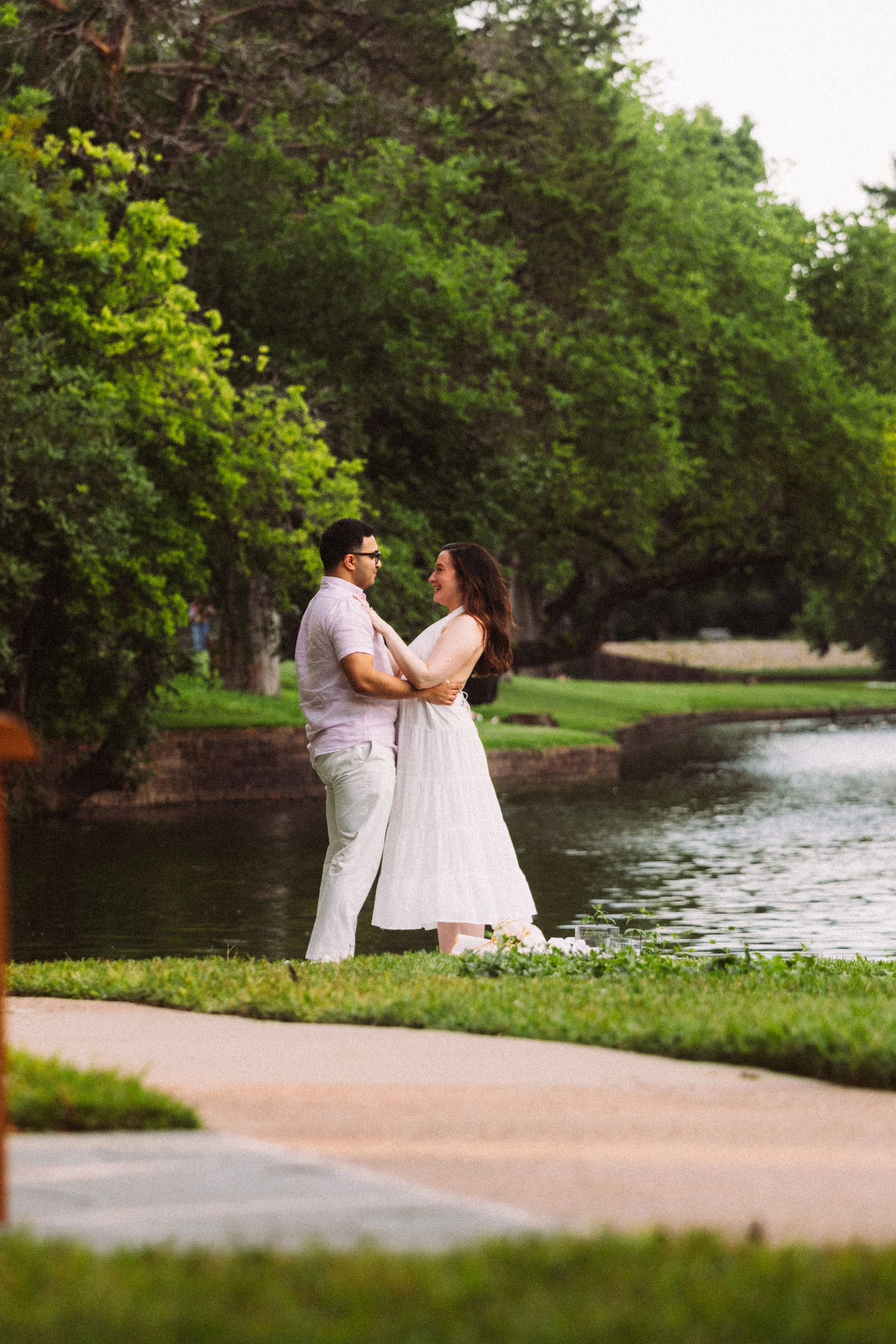 A couple stands near a lake on a grassy area, looking at each other and embracing. The woman is wearing a white dress and the man is dressed in a light-colored shirt and pants. There are trees in the background and a picnic setup with a blanket, bask