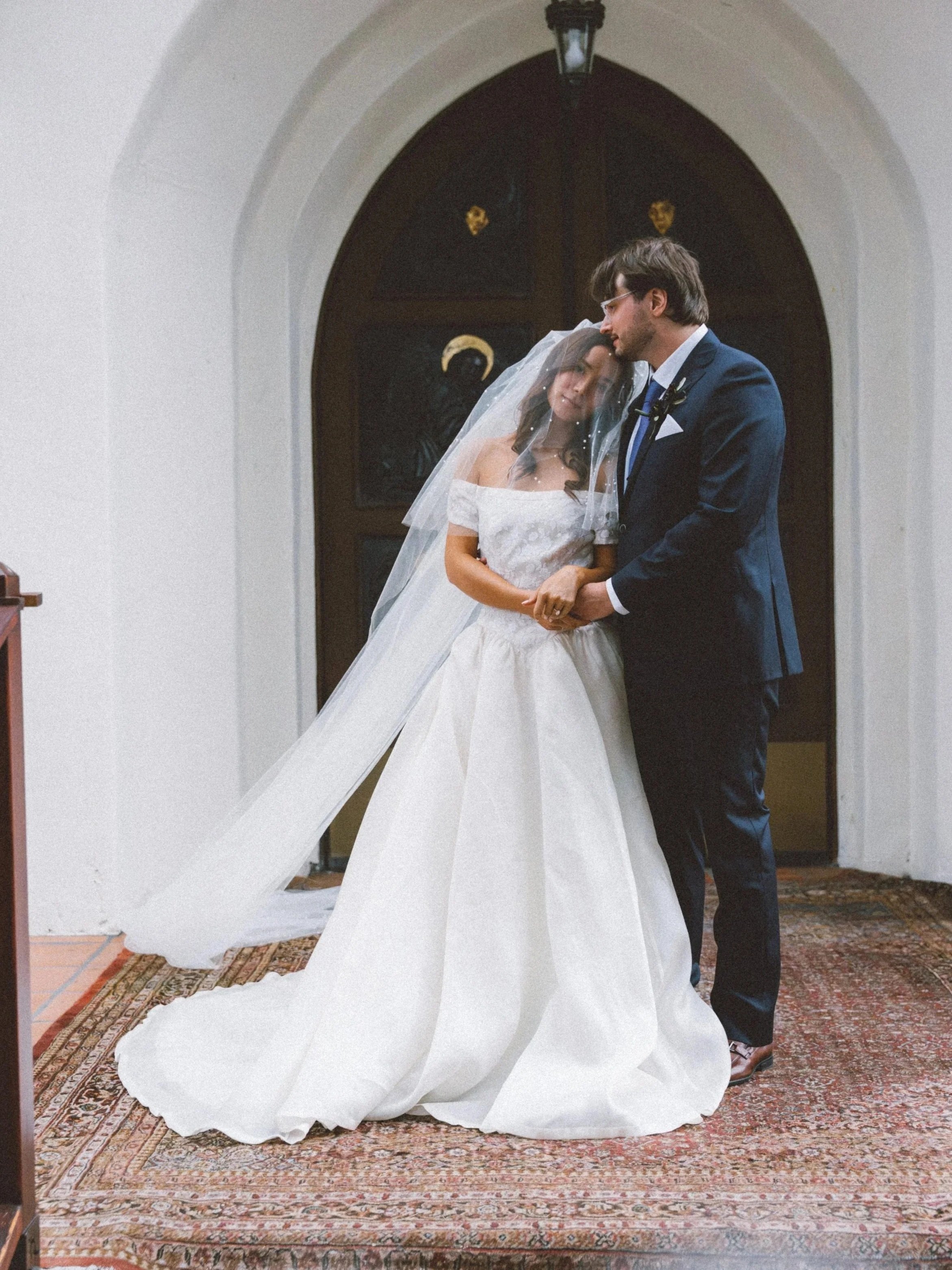 A bride and groom standing inside a church, holding hands and smiling at each other. The bride is wearing a white wedding dress and veil, and the groom is wearing a dark suit with a tie. They are standing on a patterned area rug, with a dark wooden door and a hanging lantern in the background.