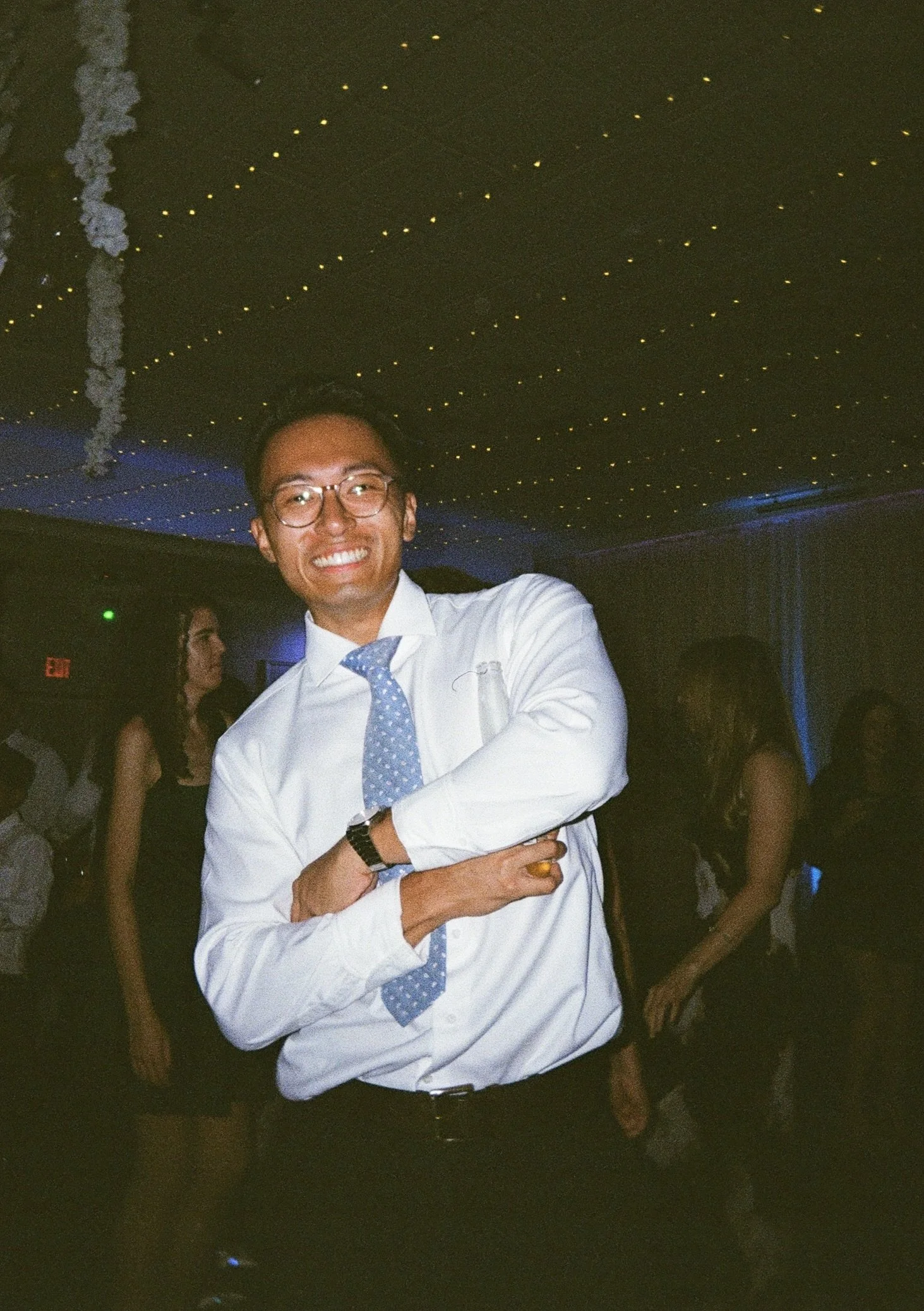 A man wearing glasses and a white dress shirt with a blue polka-dot tie smiling at a party or celebration, with a dark background decorated with small yellow lights and a ceiling with string lights.