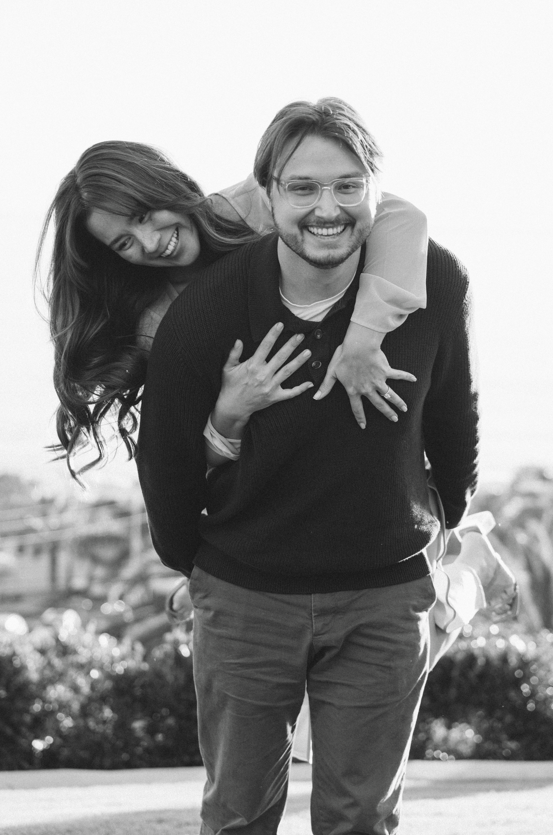 A man and woman smiling, with the woman giving a piggyback ride on the man's back, in an outdoor setting.