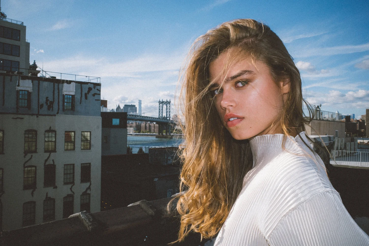 A young woman with long, wavy hair standing on a city rooftop with a view of the Brooklyn Bridge and Manhattan skyline in the background on a clear day.