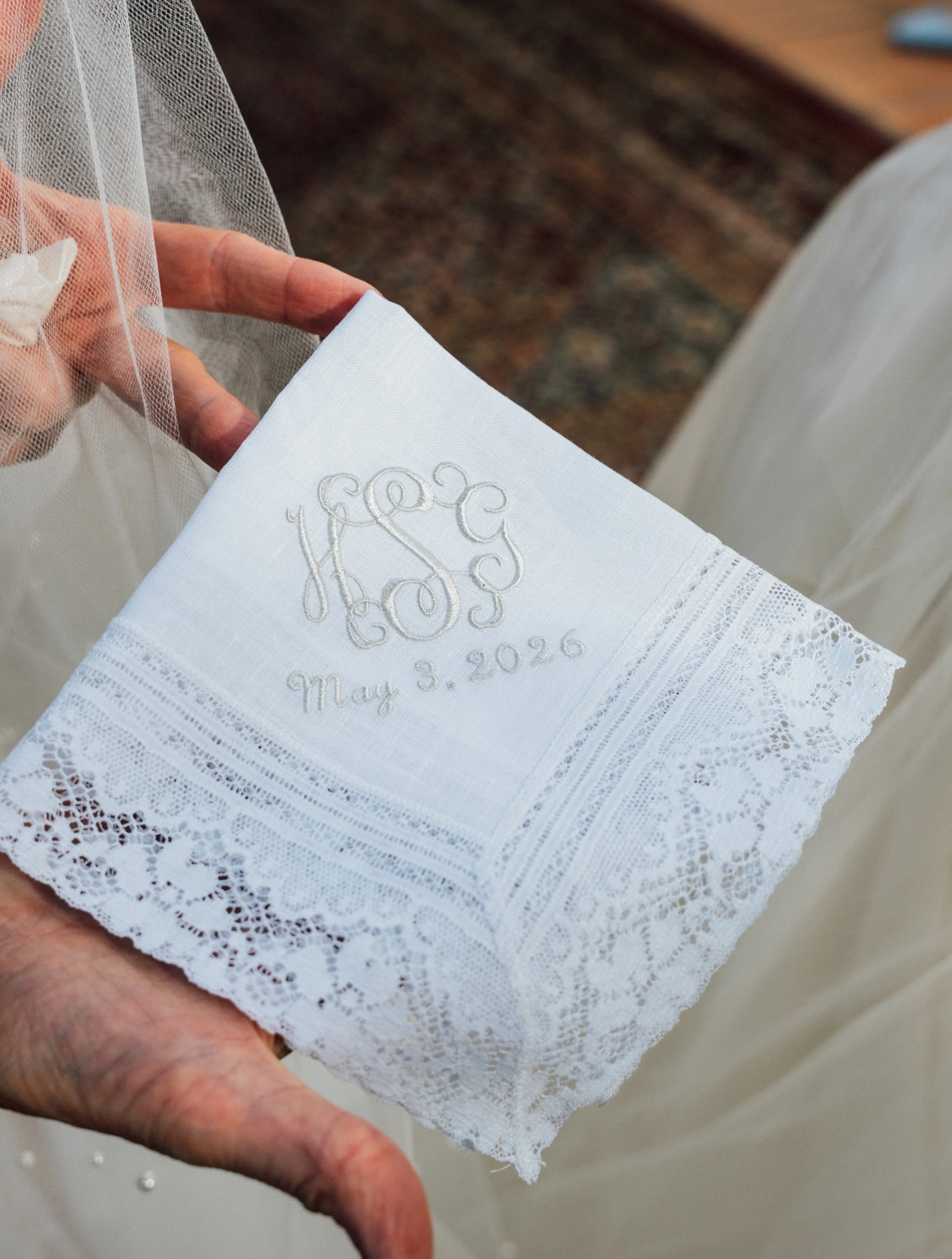 Close-up of a hand holding a white embroidered hand towel with lace edges, the towel has monogrammed initials and a date, with a lace veil in the background.