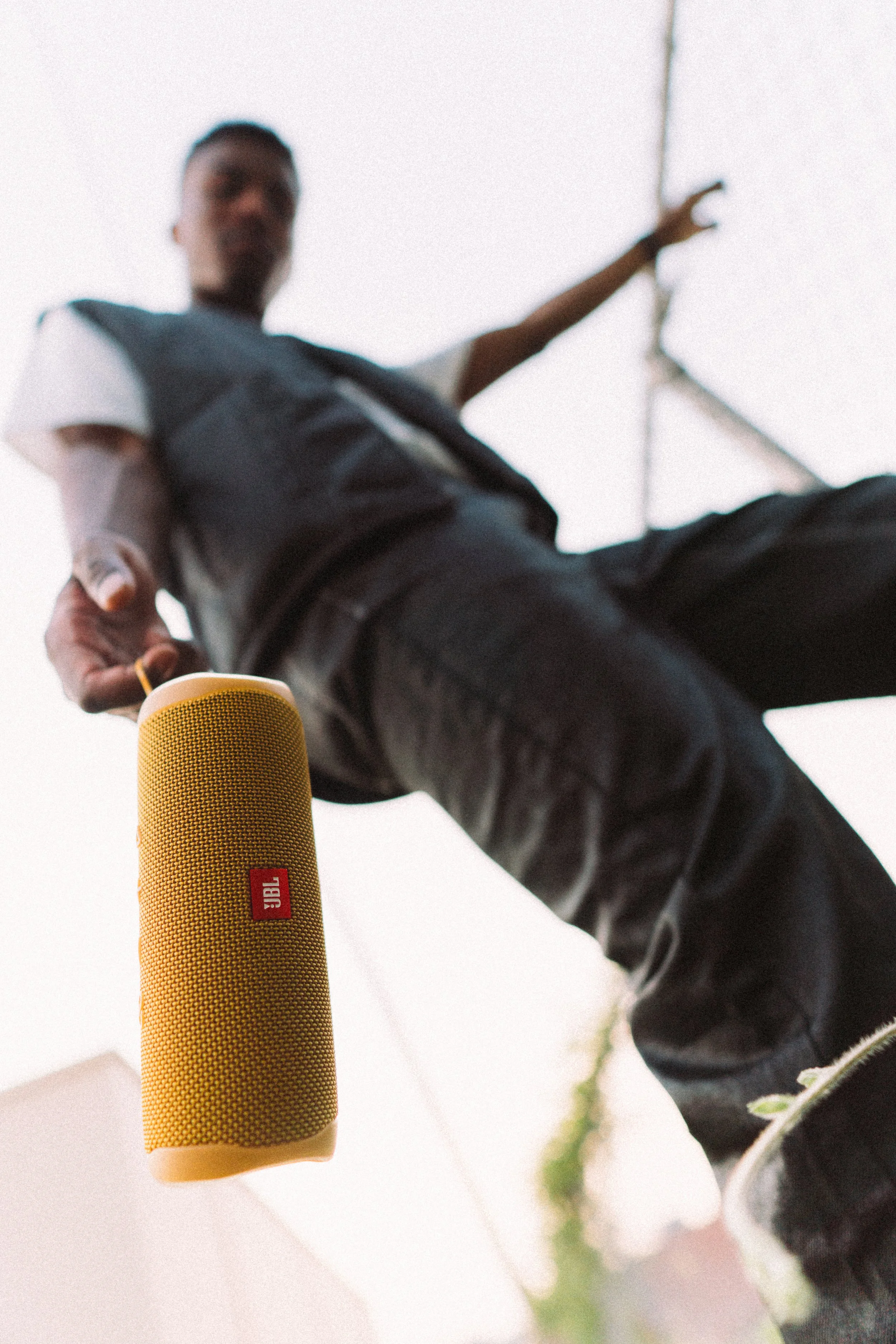 Low-angle view of a person holding a yellow JBL portable speaker, with a finger pointing, against a bright sky background.