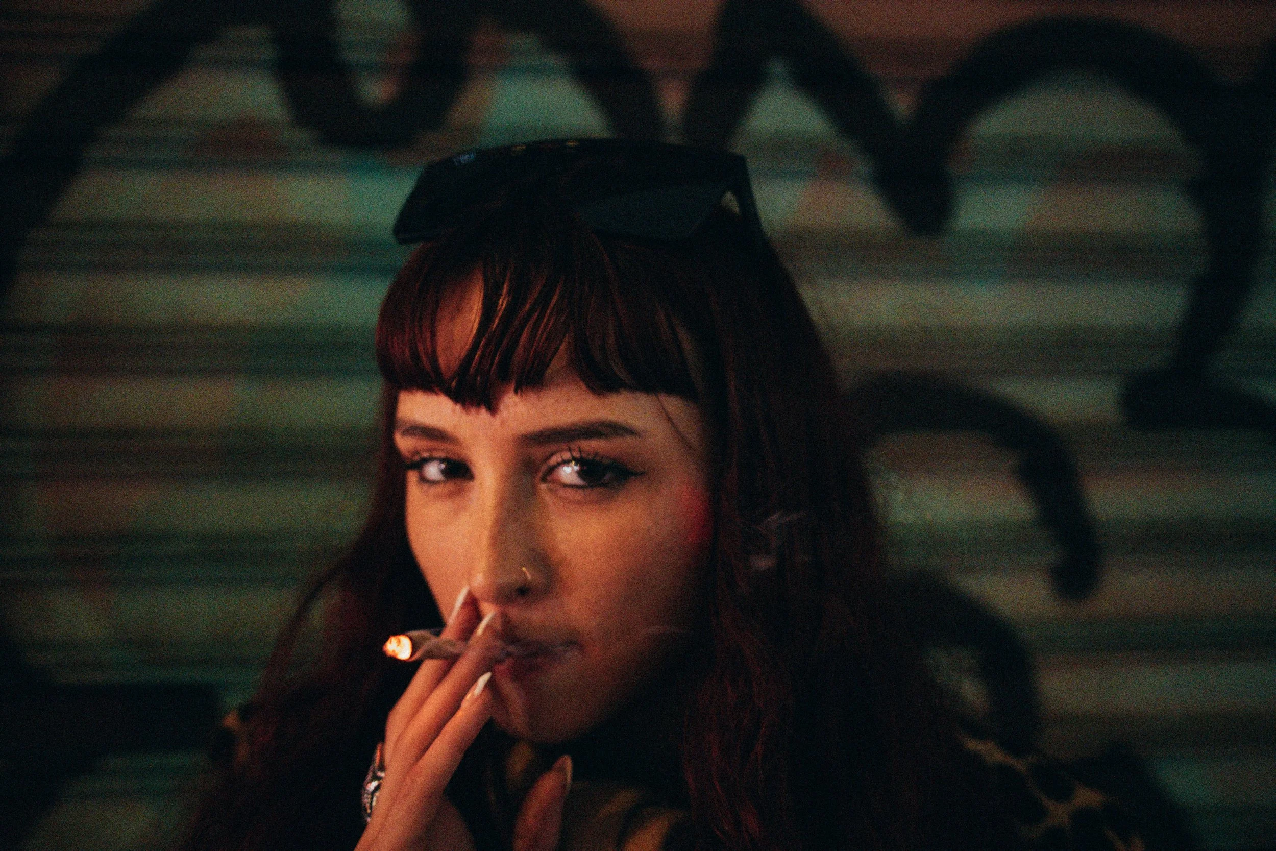 A woman with dark hair and bangs smoking a cigarette in a dimly lit setting, with her eyes partially visible.