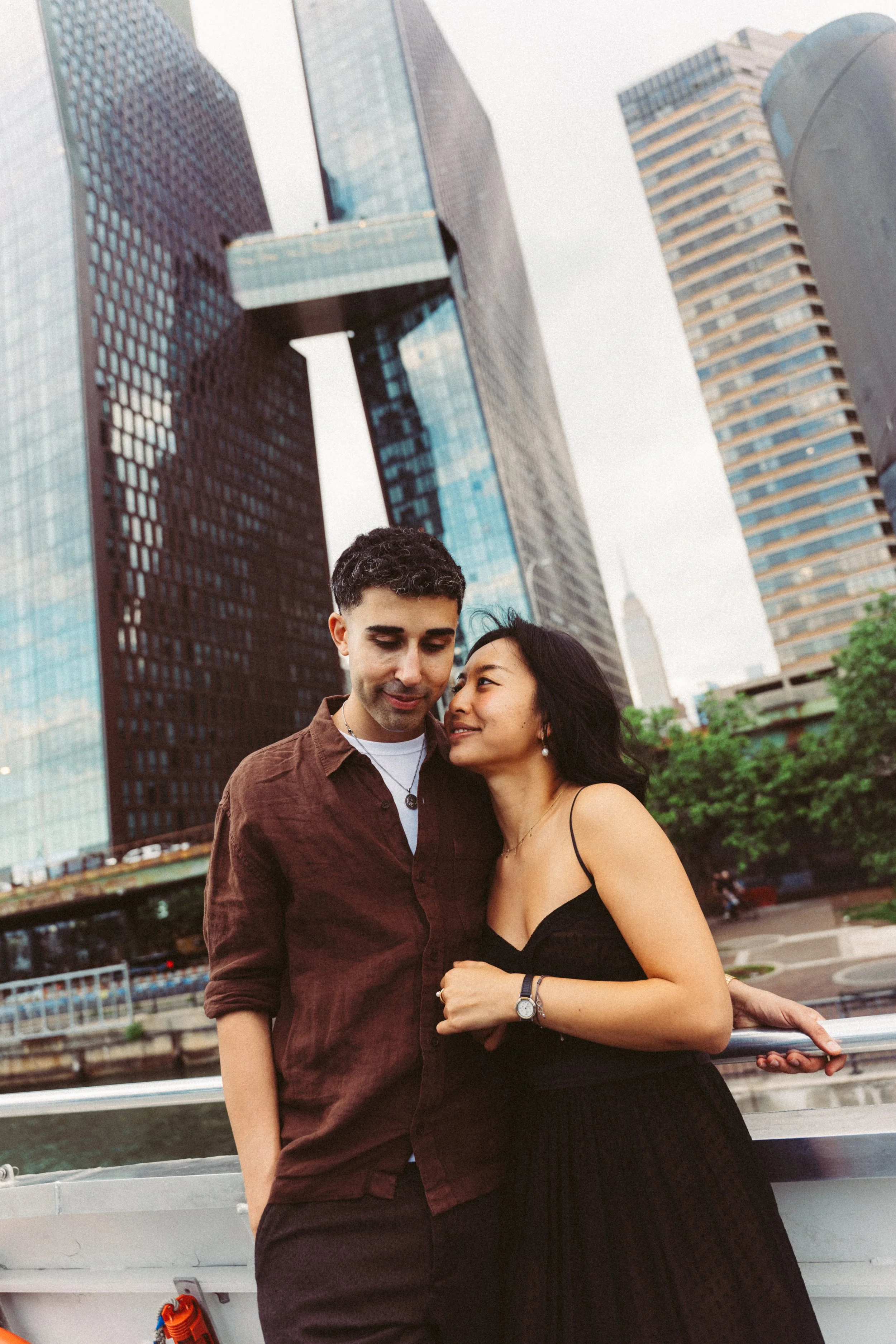 A young couple stands close together on a boat with city skyscrapers in the background, smiling and looking into each other's eyes during sunset.