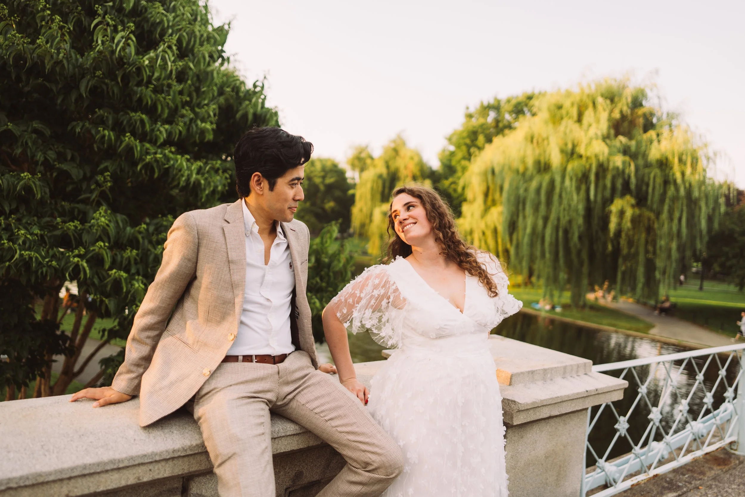A man dressed in a beige suit and a woman in a white lace dress are sitting and standing close to each other on a stone ledge in a park with lush green trees and a pond in the background during sunset.