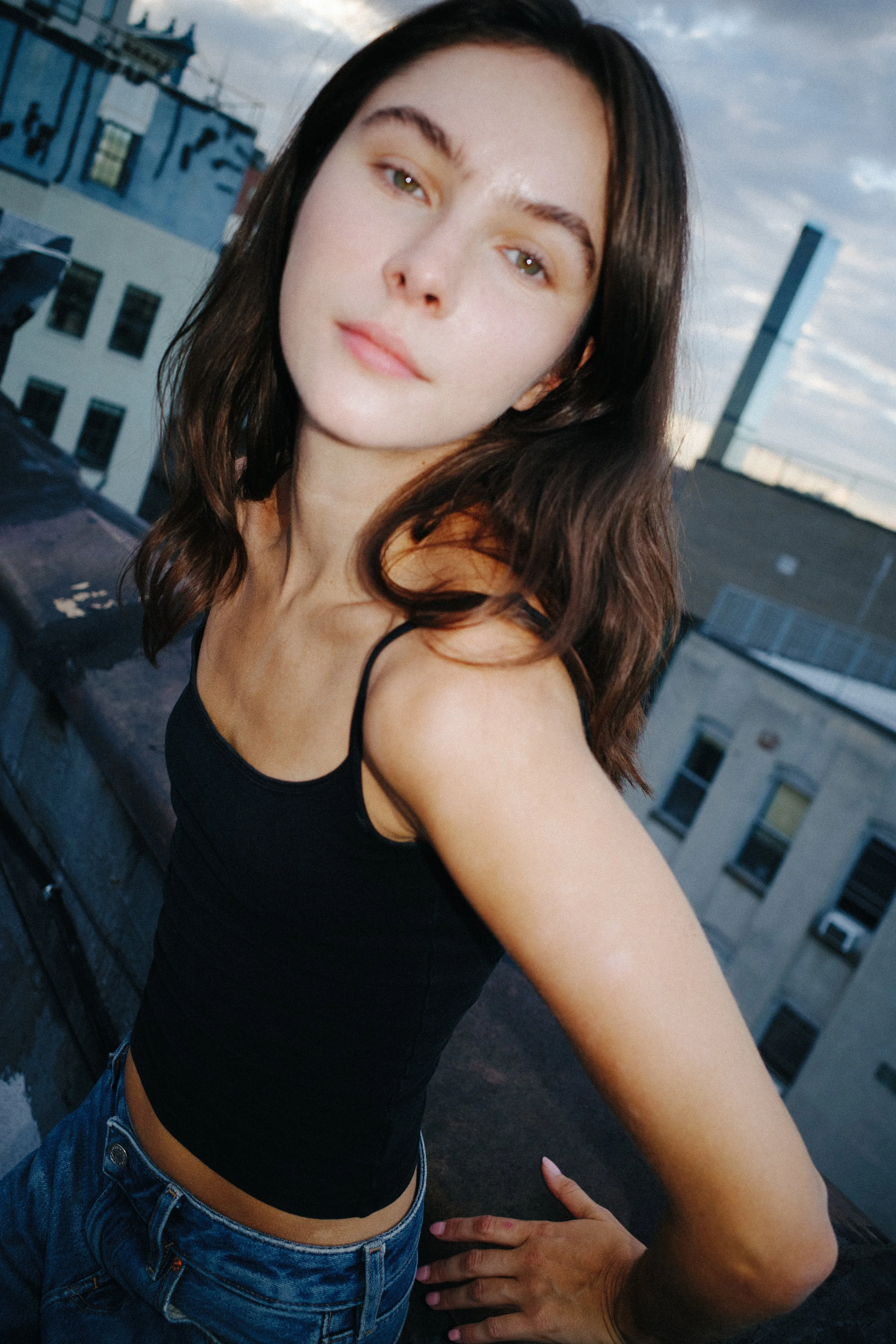 A young woman taking a selfie on a rooftop with city buildings in the background during the evening.