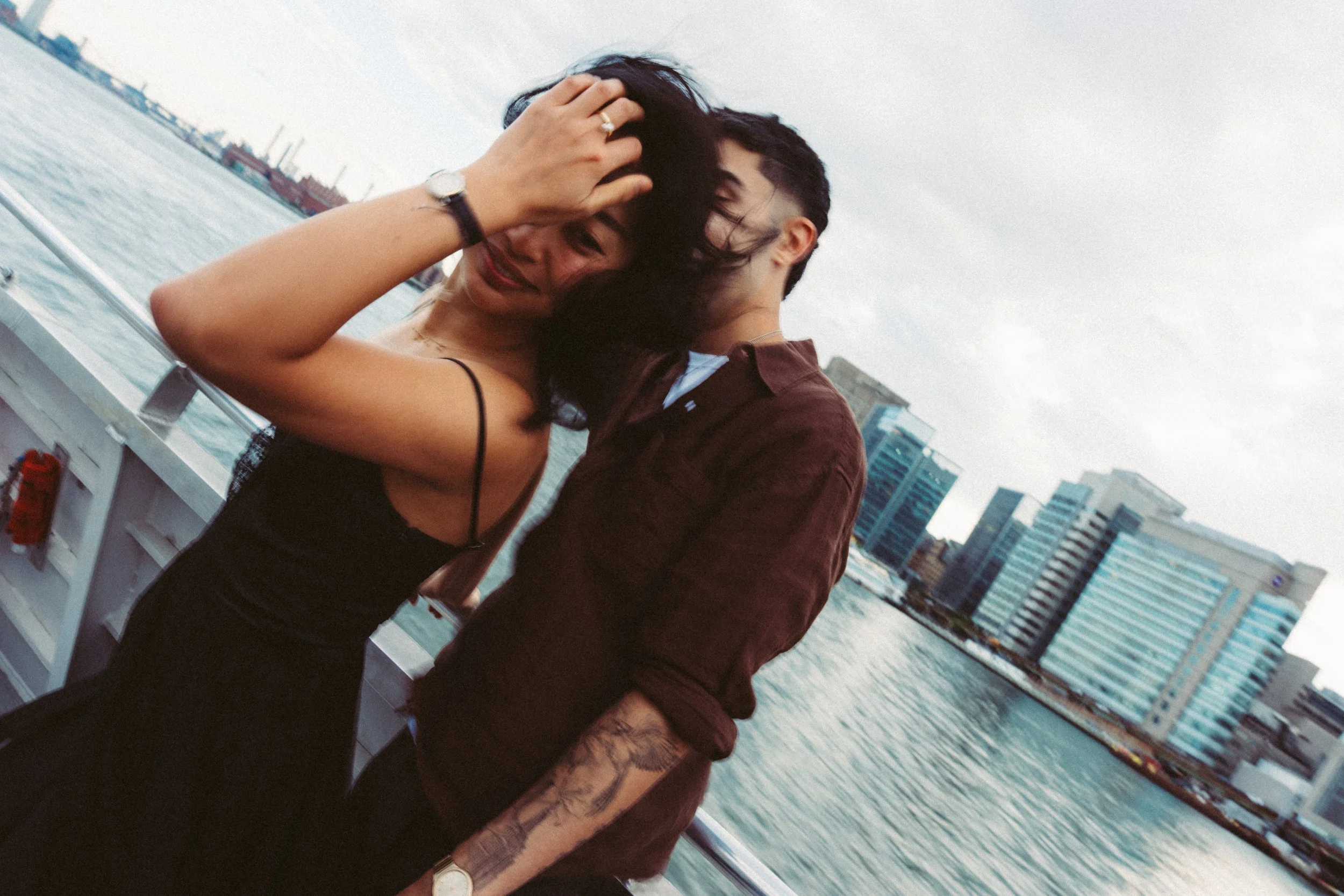 A woman and a man on a boat with a city skyline in the background. The woman has long dark hair and is wearing a black sleeveless top, and the man has short dark hair, a beard, and is wearing a brown jacket. They are embracing while smiling, with the