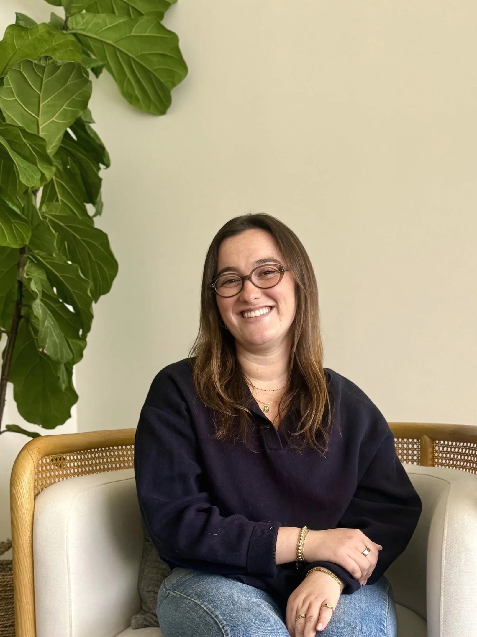 A person with long hair sitting on a brown leather chair, wearing a beige cardigan and black pants, with a plant in the background.