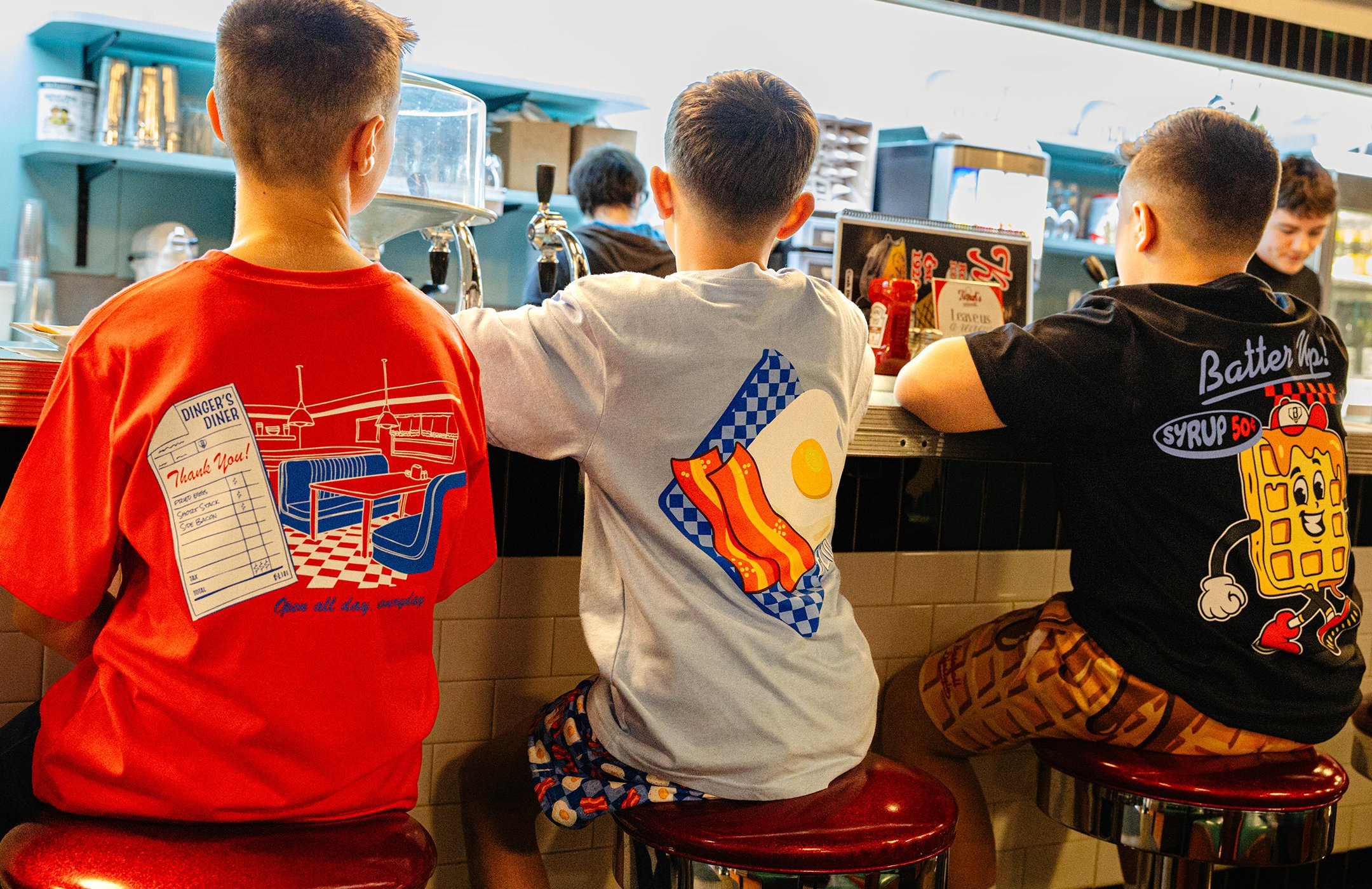 Three boys sitting at a counter in a diner, viewed from behind. They are wearing colorful Baseball Lifestyle 101 t-shirts with graphic prints. The area behind the counter has various kitchen items and a person preparing food.
