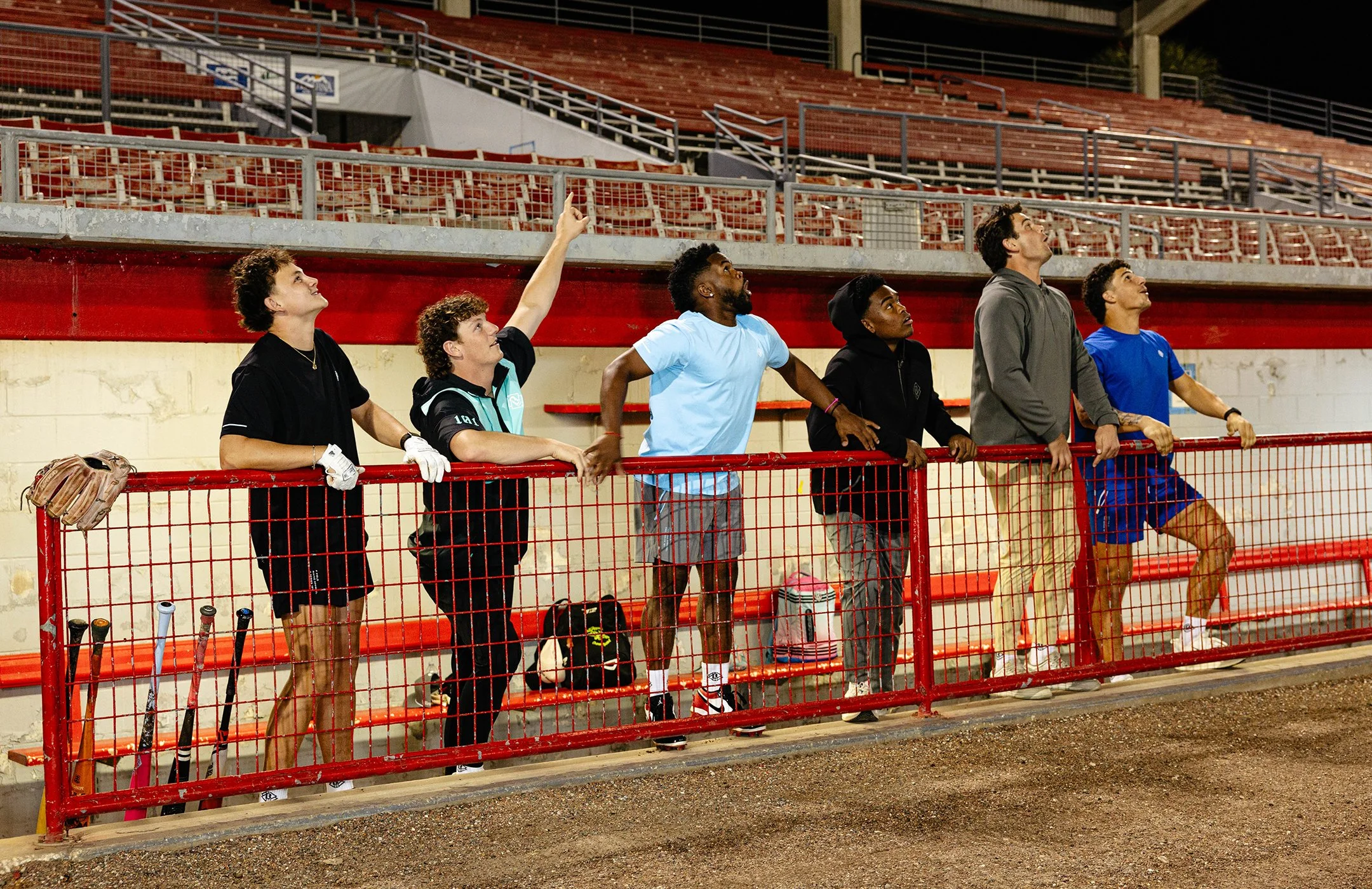 A group of six young male athletes watching and pointing at something above at a sports stadium’s dugout area at night. They are wearing Baseball Lifestyle 101 athletic clothes.
