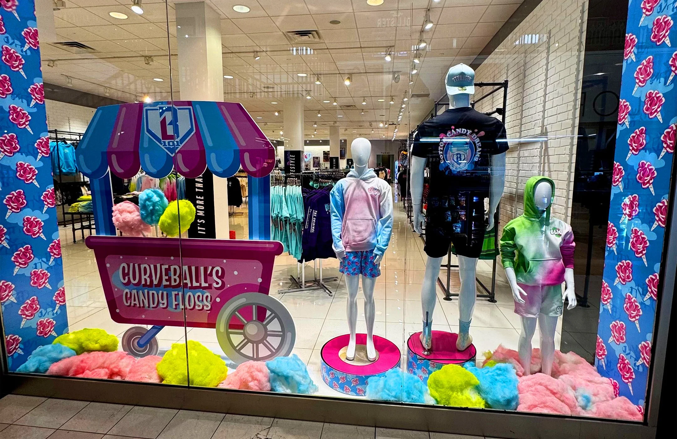 Store display window featuring mannequins dressed in colorful Baseball Lifestyle 101 clothing, including hoodies and shorts, with a cotton candy cart and fluffy pink, blue, green, and yellow cotton candy decorations inside the window.