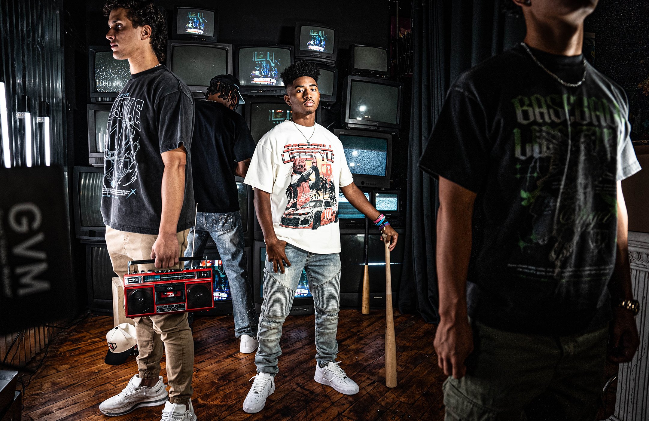 Four young men in a room with multiple TV screens, some turned on and some off, with vintage video game graphics on the screens. They are dressed in streetwear Baseball Lifestyle 101, with one holding a boombox, and another with a baseball bat.