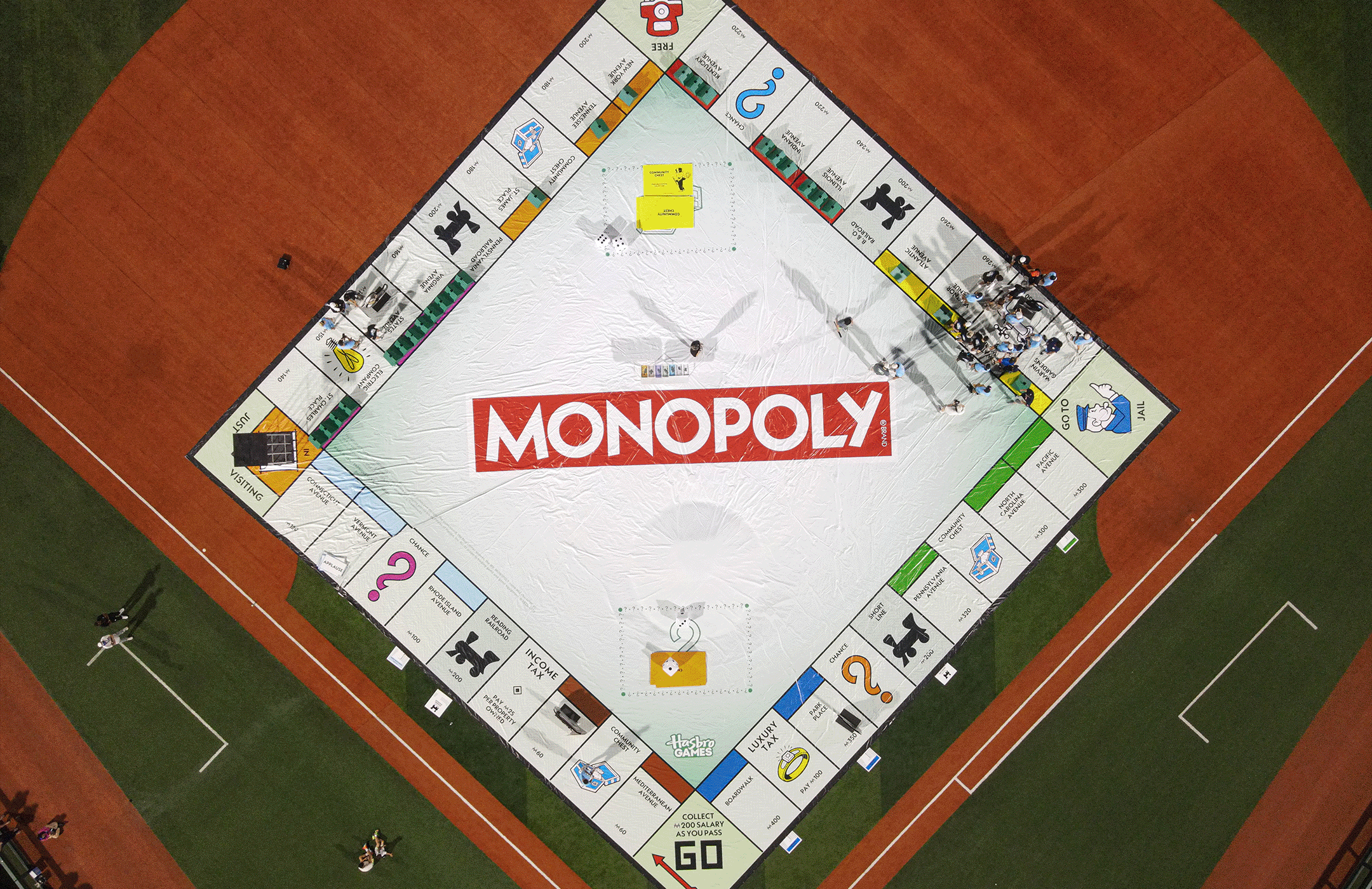 An aerial view of a 90 foot by 90 foot Monopoly board game set up on a baseball field. The game board is square with a central white area featuring the red Monopoly logo. There are people standing and walking on and around the board.