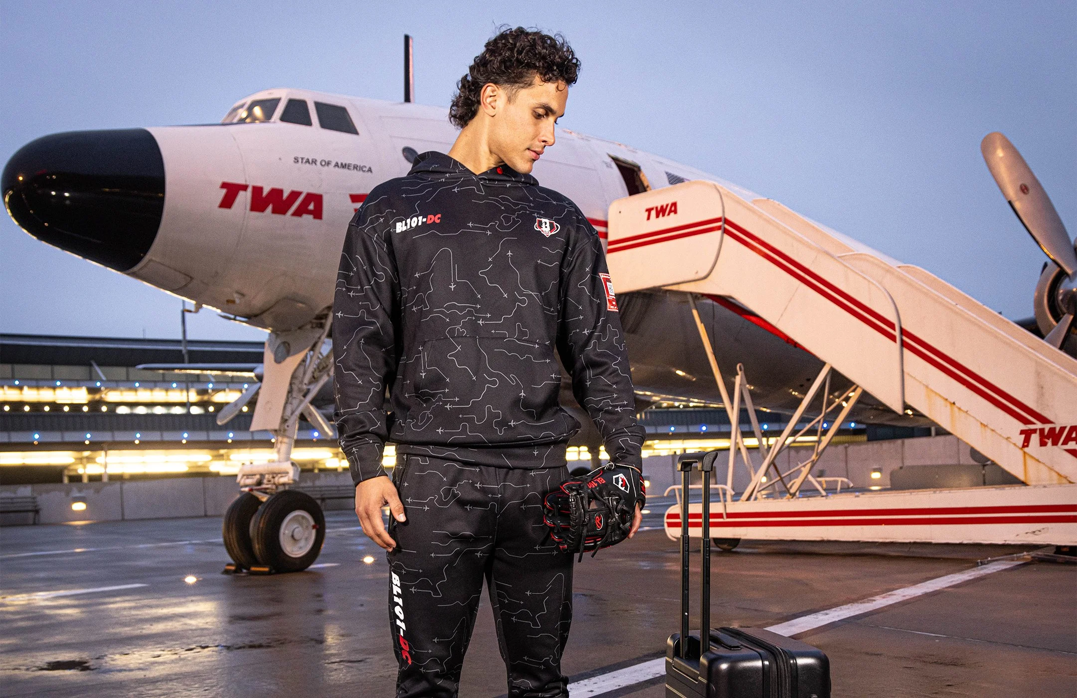 Young male model in Baseball Lifestyle 101 sweatsuit, holding his flight gloves and luggage on an airport tarmac at dusk, with a retro-style airplane in the background.