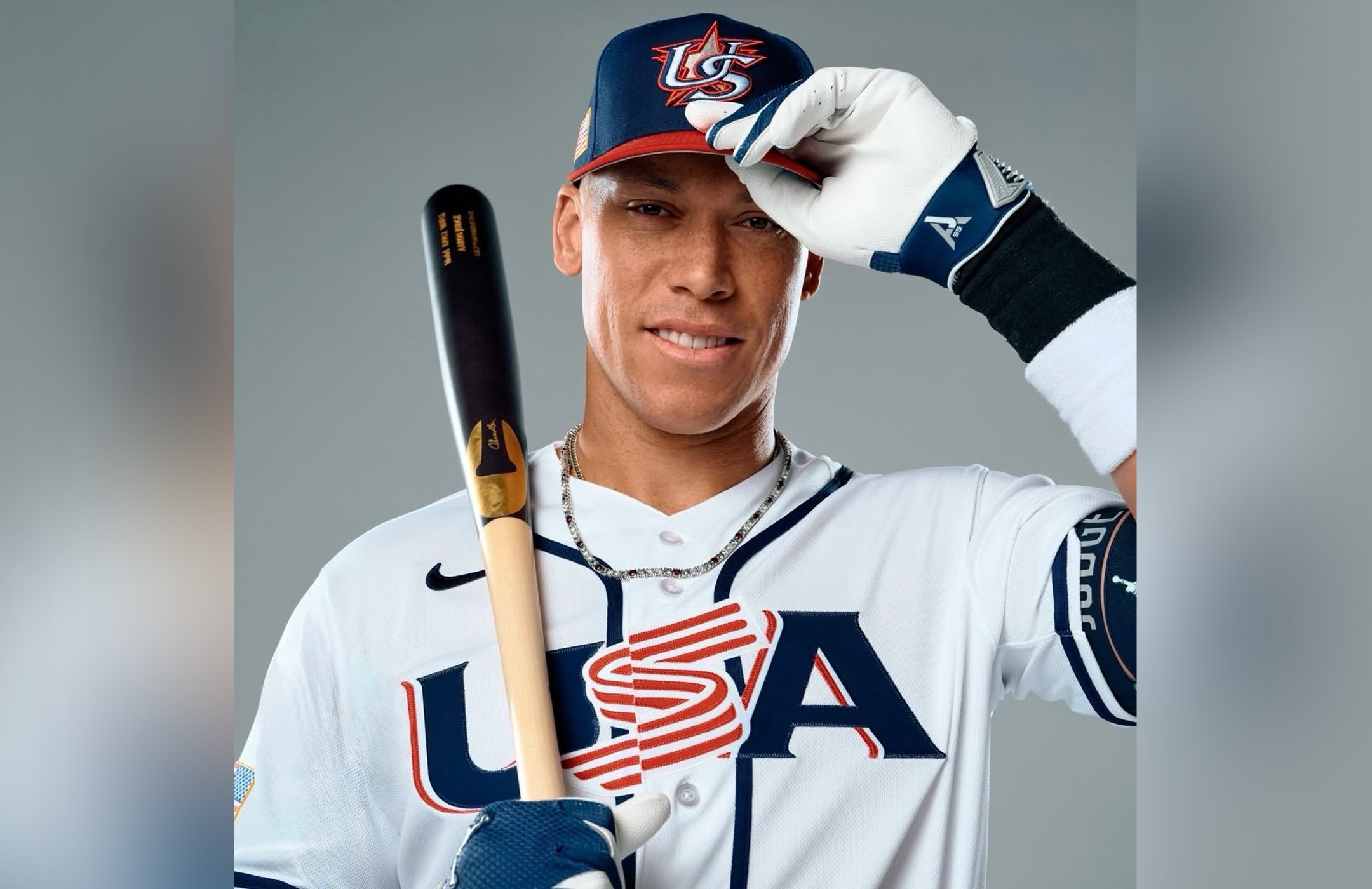 New York Yankees Aaron Judge in a USA team uniform holding a bat, wearing a USA cap and white batting gloves, and smiling at the camera. AJ99 logo featured on his batting glove wrist.