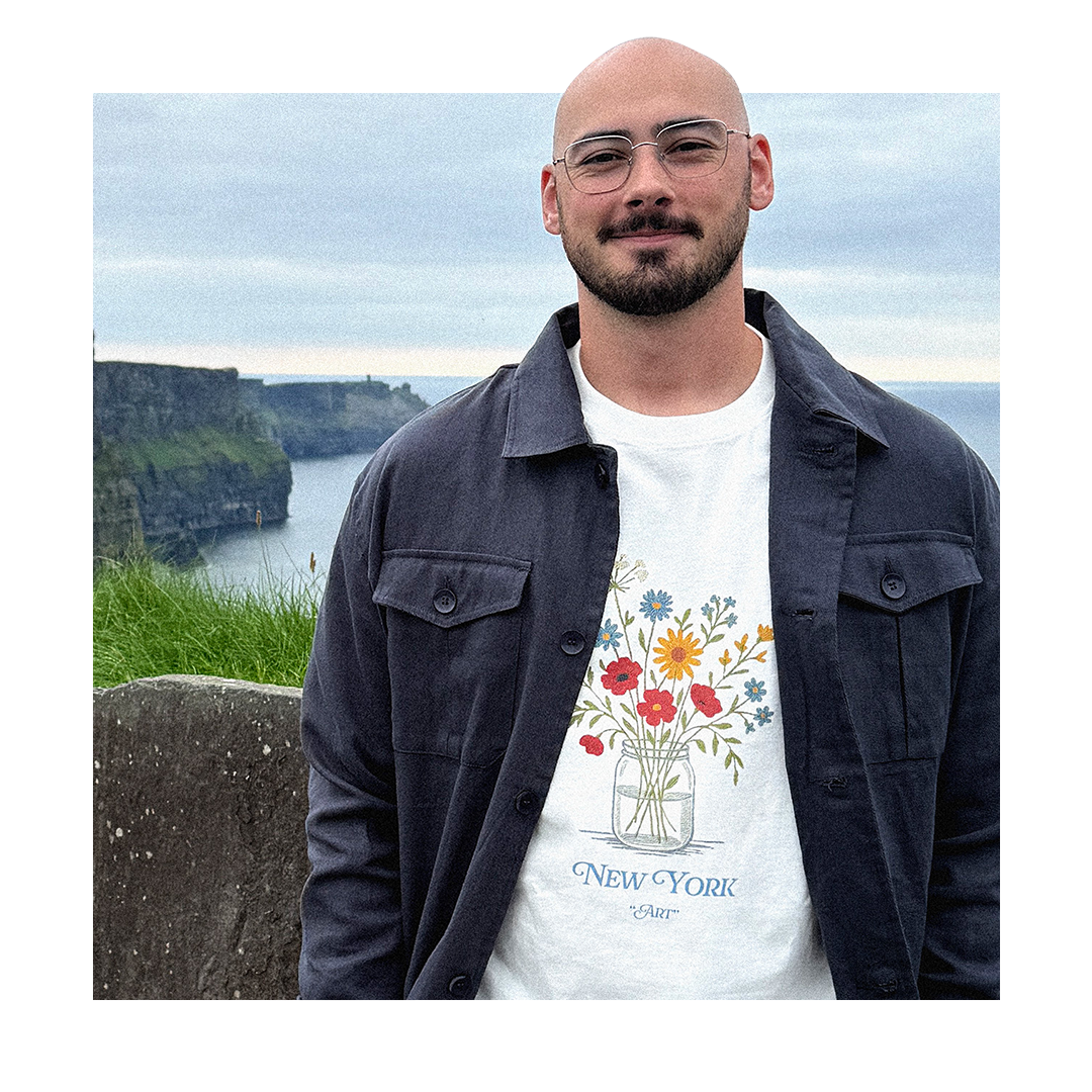 Colin Casaccio standing over the Cliffs of Moher in Ireland, wearing a white T-shirt with flower illustrations and text 'New York Art' under a dark jacket.