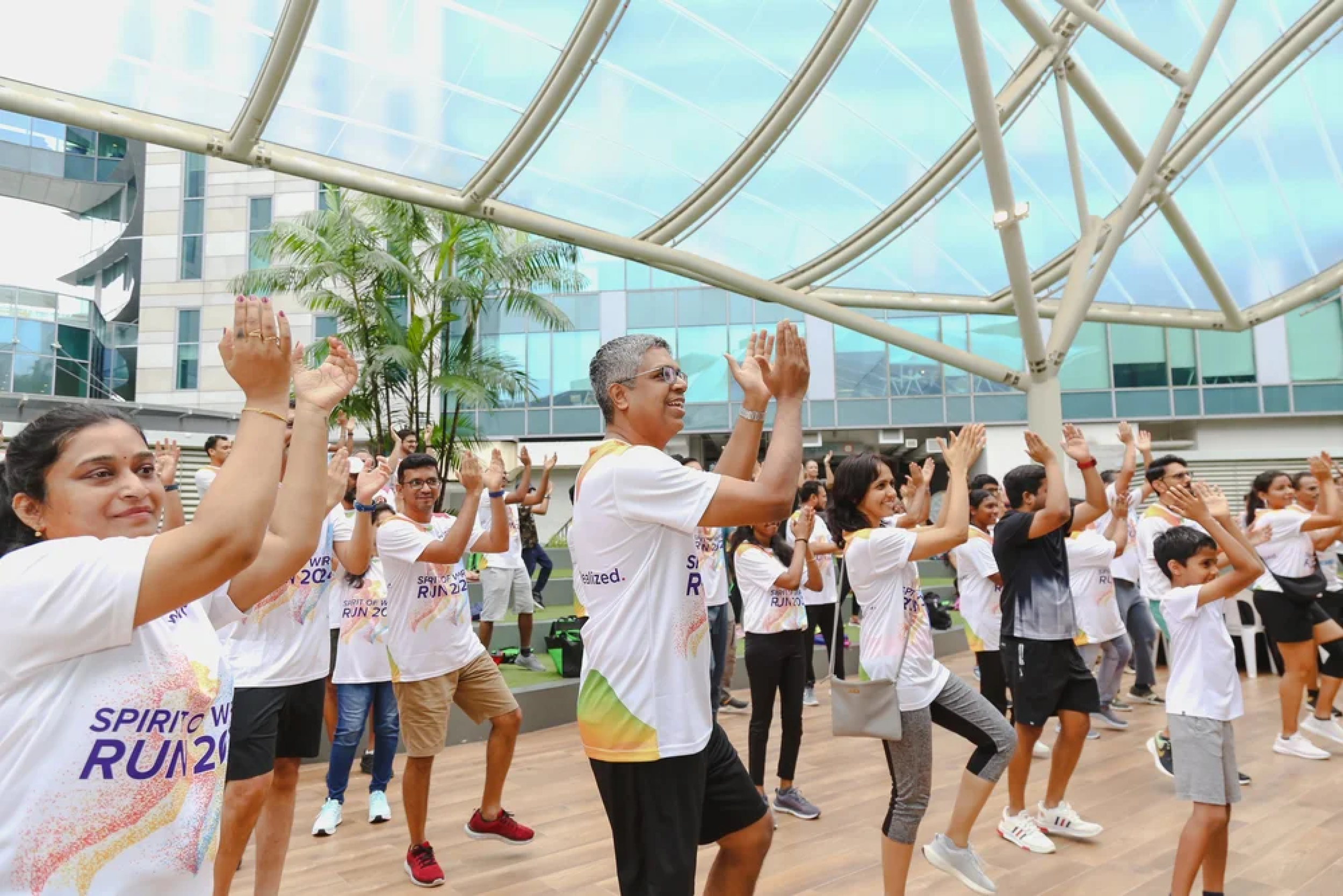 A group of diverse people participating in a group exercise or dance event indoors, with a modern glass and steel roof and windows, and some indoor plants in the background.