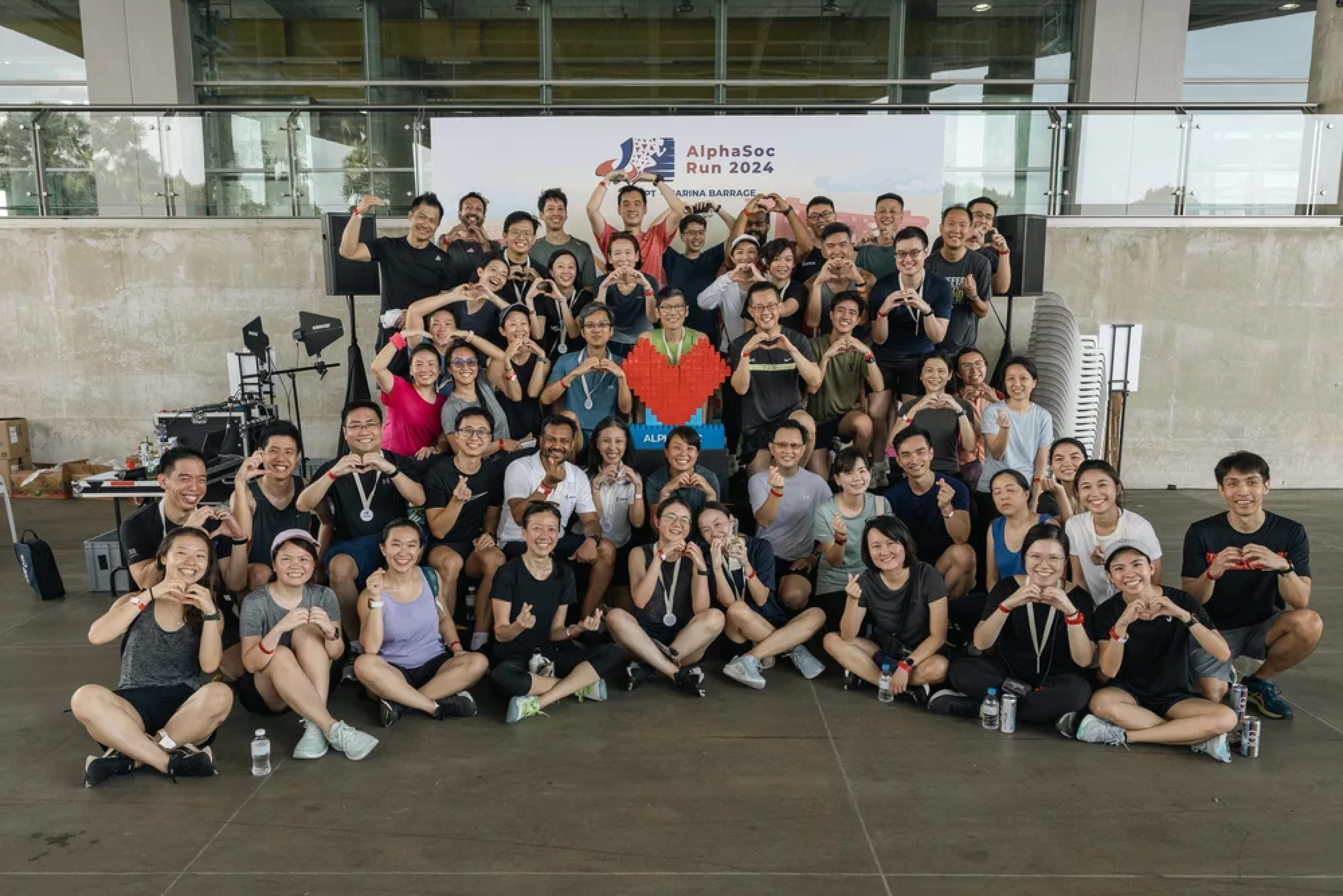 Group of people gathered indoors, smiling and making heart signs with their hands, at the AlphaSoc Run 2024 event with a large banner in the background.