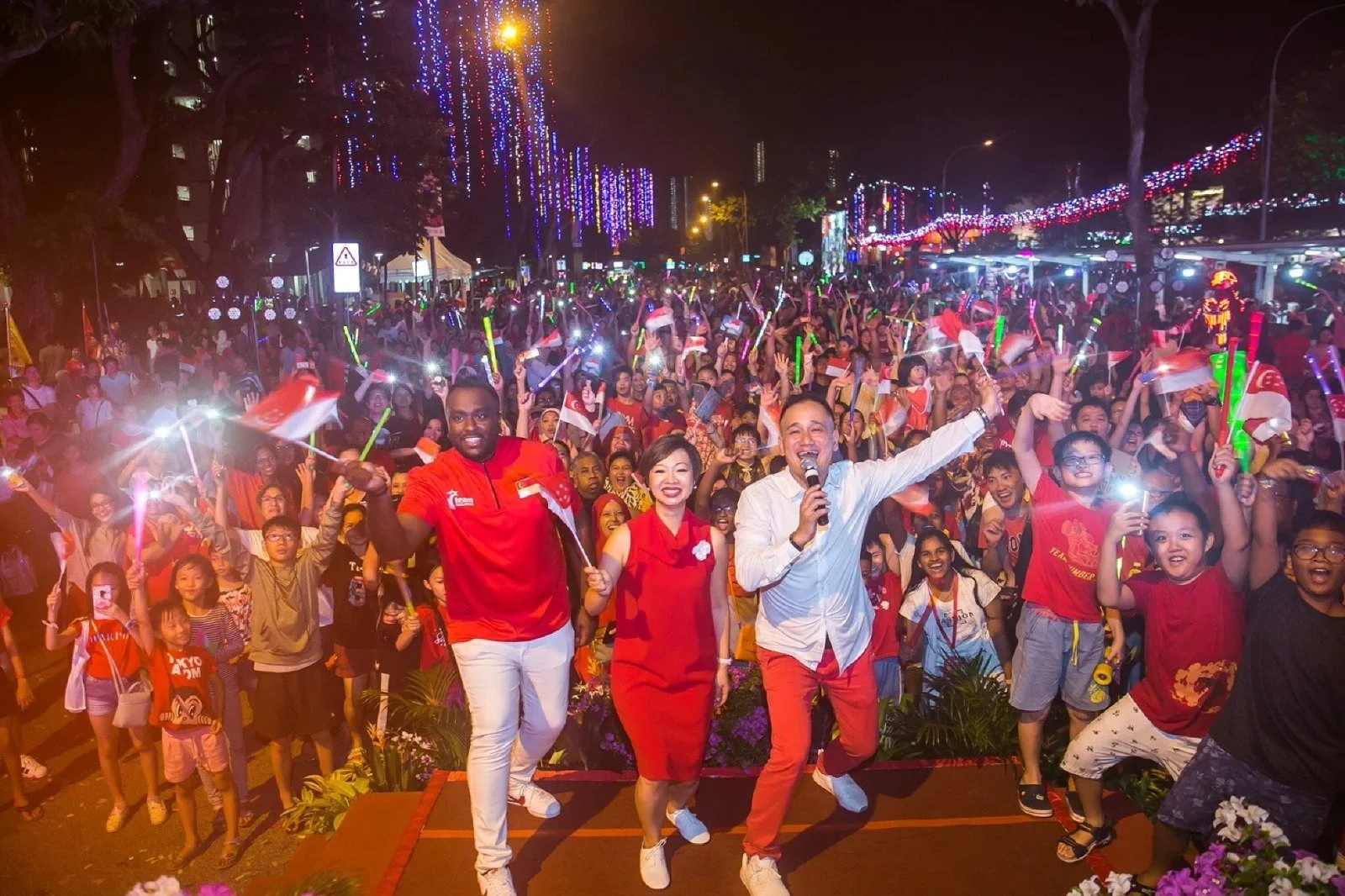 A large crowd of people celebrating at a nighttime event, many holding flags and glow sticks, with three people in the foreground smiling and waving at the camera, surrounded by colorful lights and decorations.