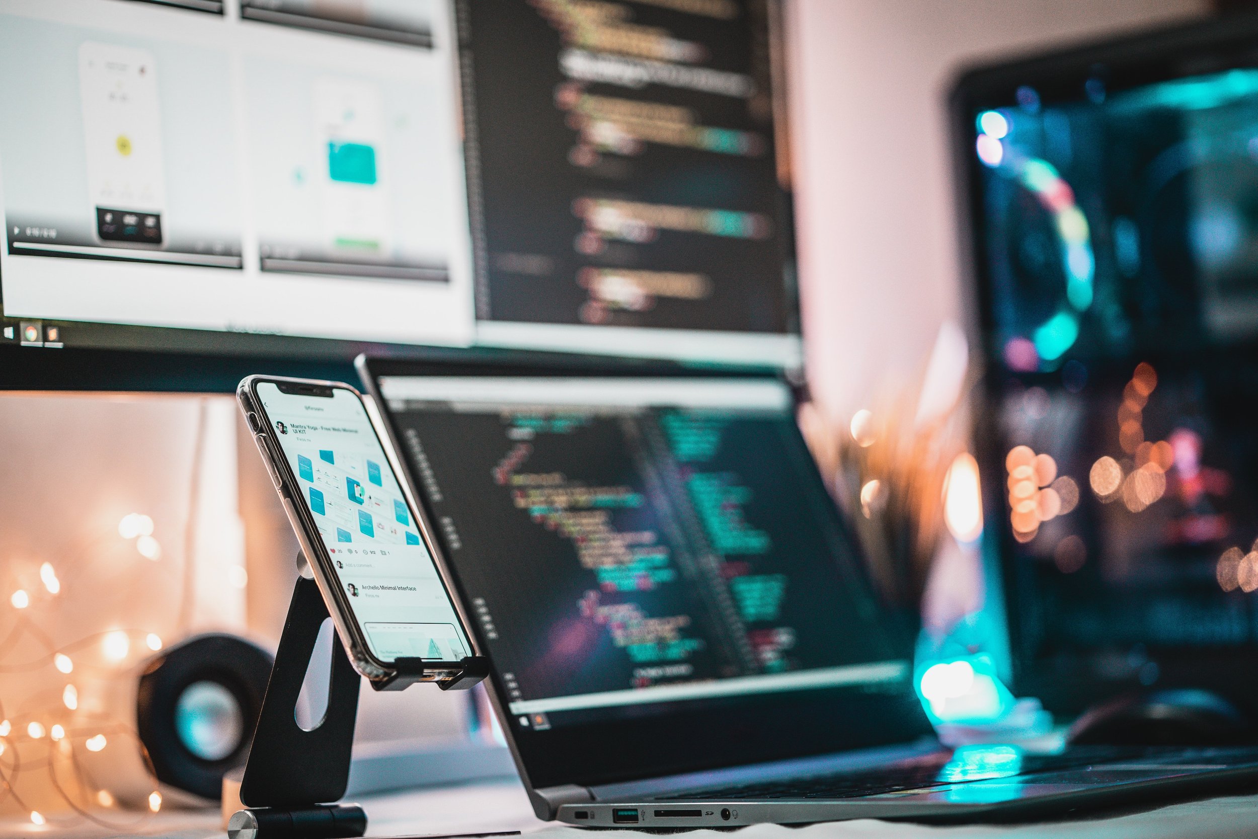 A workspace with a computer monitor displaying code, a smartphone on a stand showing an app, and blurred colorful bokeh lights in the background.