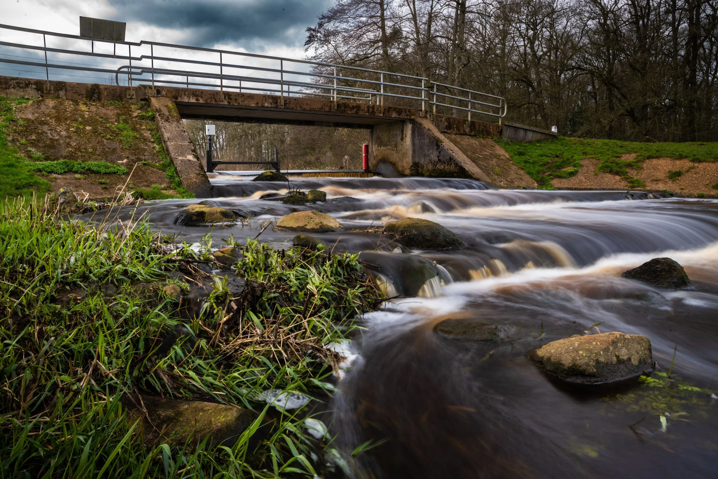 Stream flowing under a stone bridge with greenery on the riverbank and trees in the background.