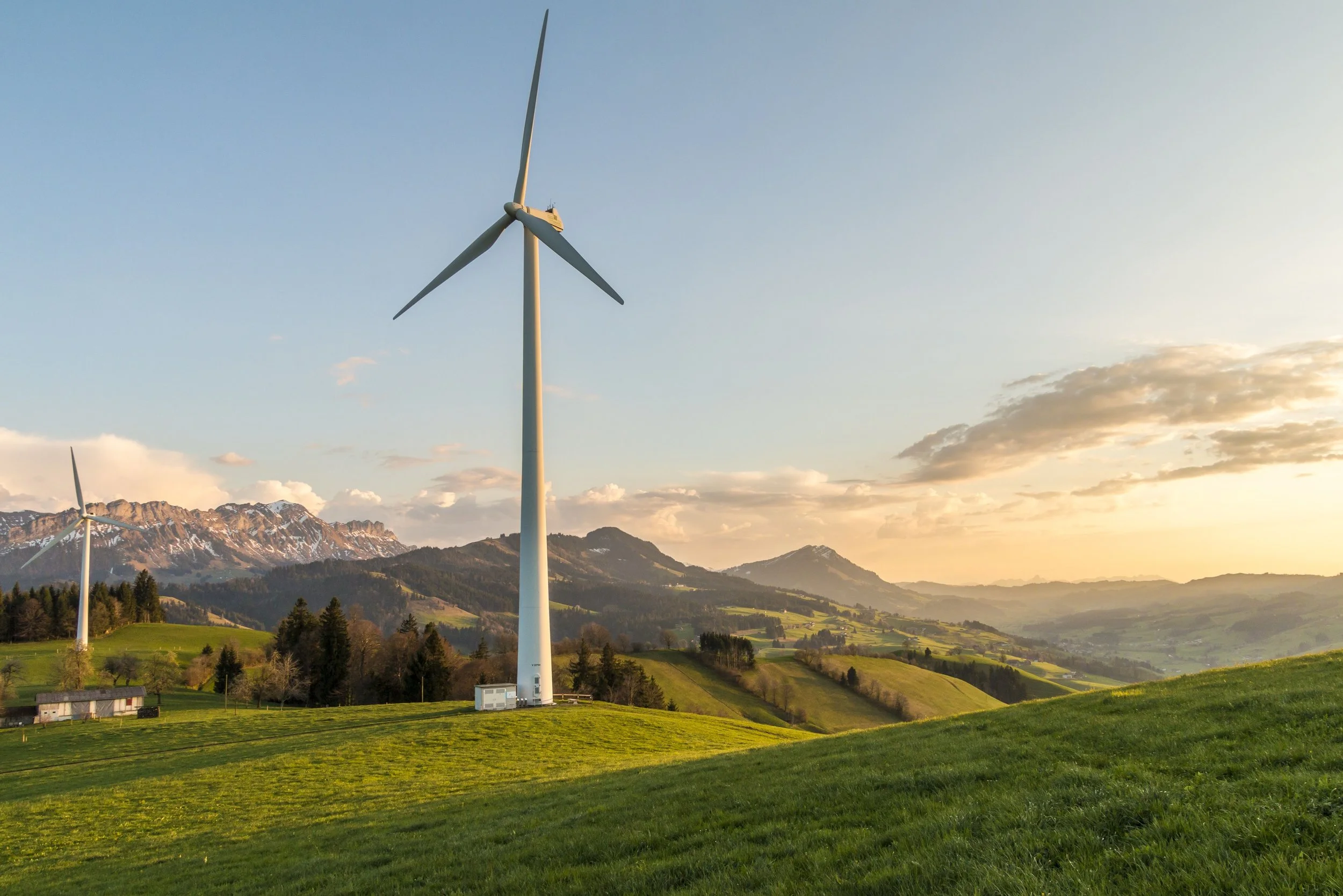 Landscape with wind turbines on green rolling hills, snow-capped mountains in the background, and a partly cloudy sky at sunset.