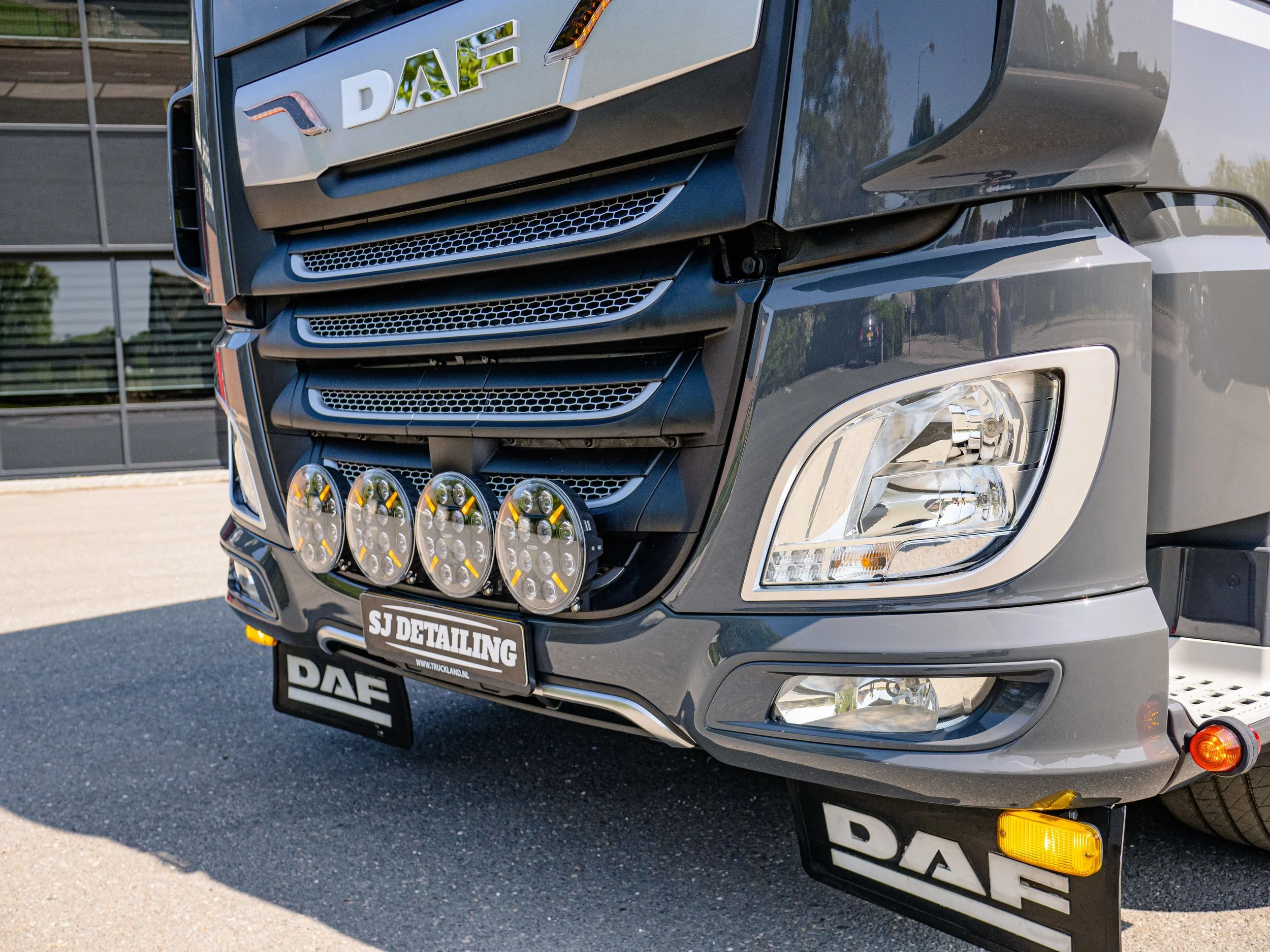 The front of a black DAF semi-truck with multiple off-road lights mounted on the bumper, parked outdoors near a modern building with reflective glass windows.