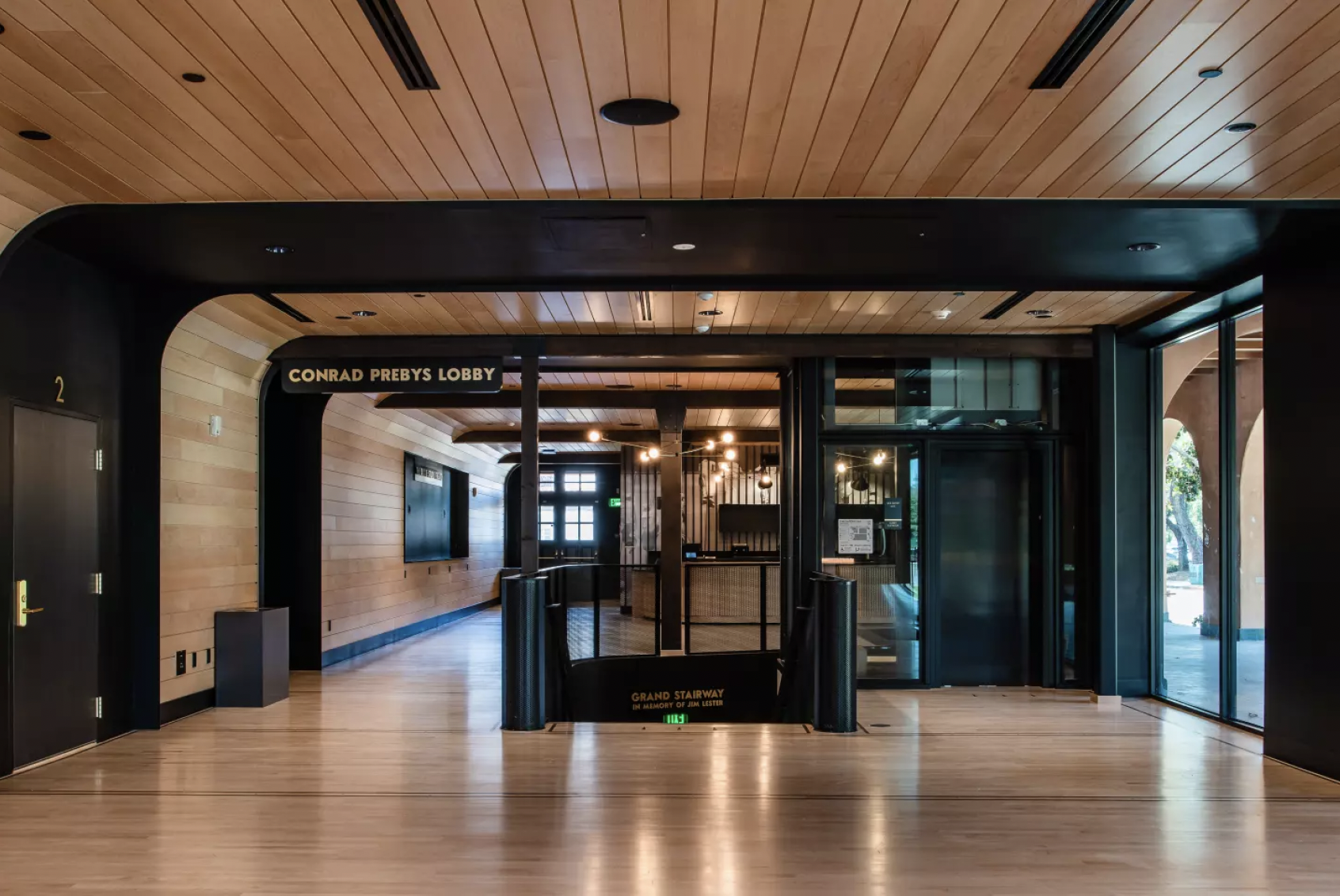 Interior of a modern building lobby with wooden ceiling and flooring, sign reading 'Conrad Prebys Lobby', and a staircase leading downward labeled 'Grand Stairway' in memory of Jim Lester.