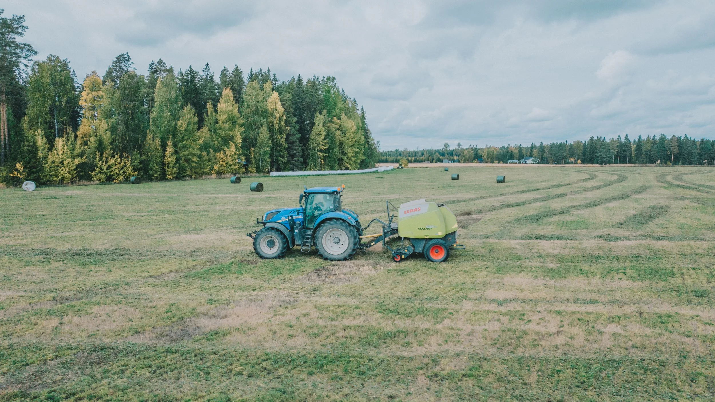 Sininen traktori pellolla vieressä paalain, ympärillä metsää ja heinärullia.