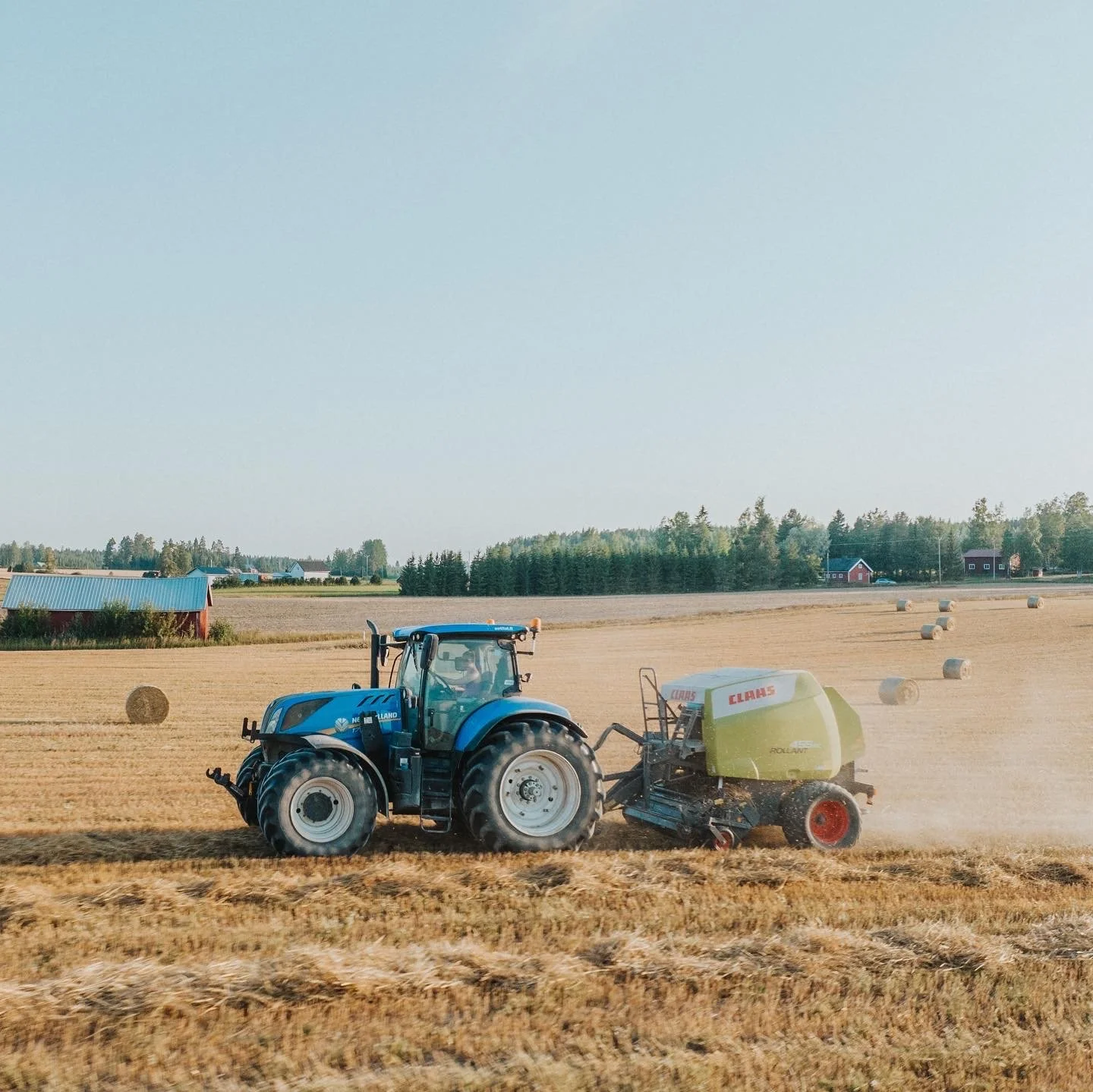 Sininen traktori pellolla paalaamassa heinää, taustalla lato ja metsää, maaseutuympäristö lämpimänä päivänä.