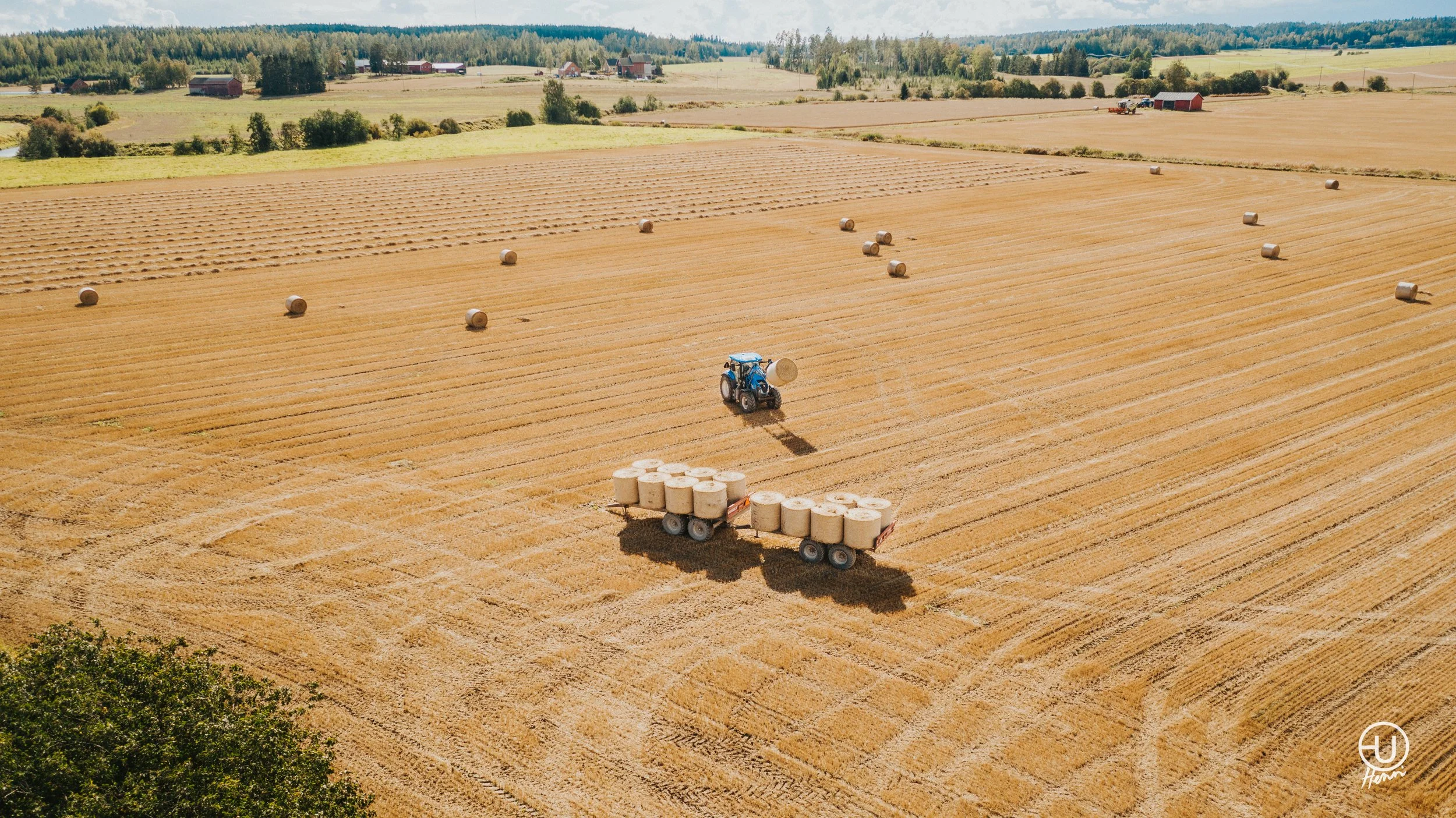 Maatilamaisema, traktorilla paaleja pellolla, taustalla metsä ja rakennuksia.