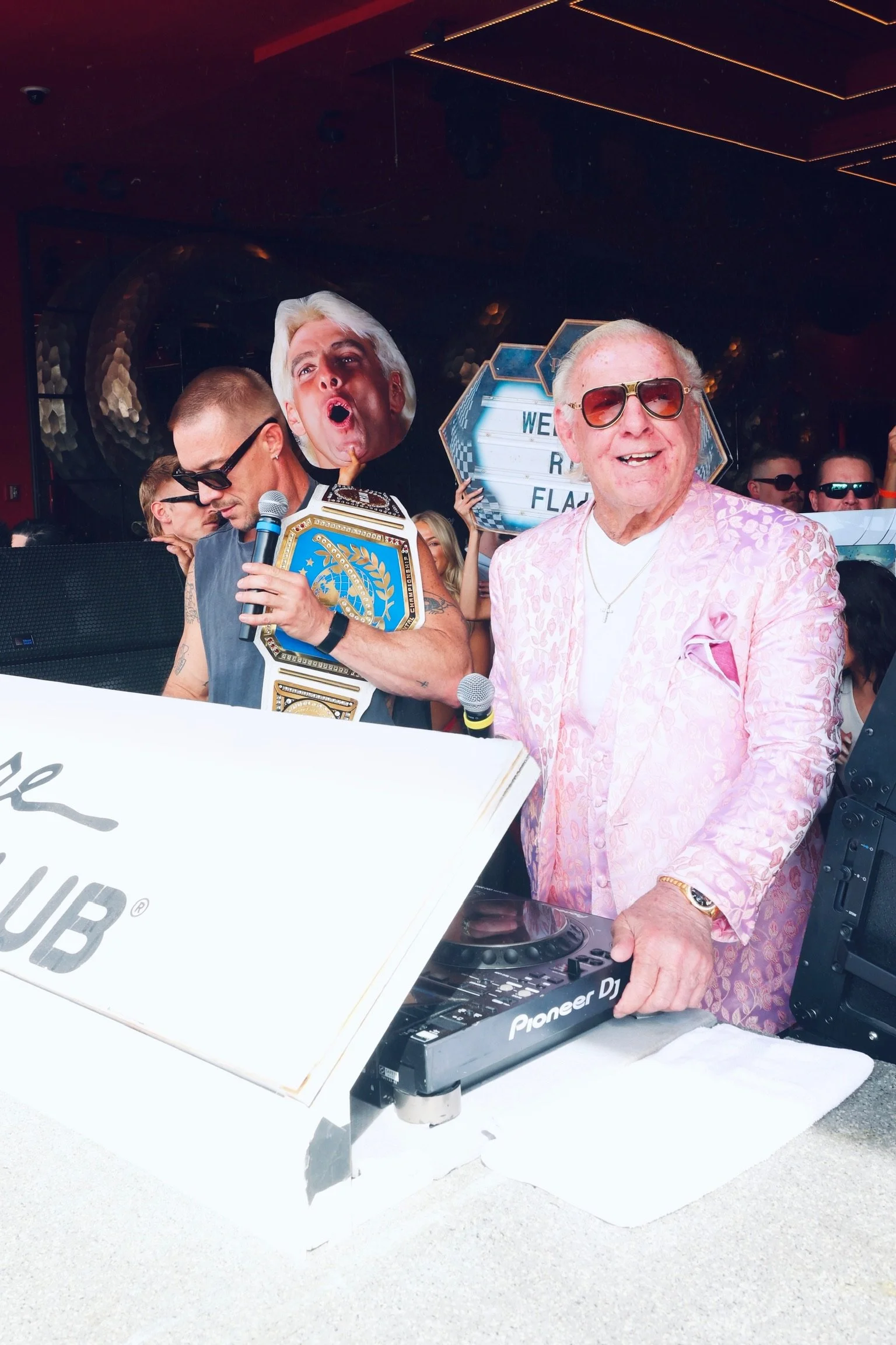  DJ Diplo stands at the Pioneer DJ decks, wearing a grey sleeveless shirt and sunglasses, looking out toward the crowd with a focused expression. In the background, Ric Flair, dressed in his signature pink patterned blazer and sunglasses, smiles broa