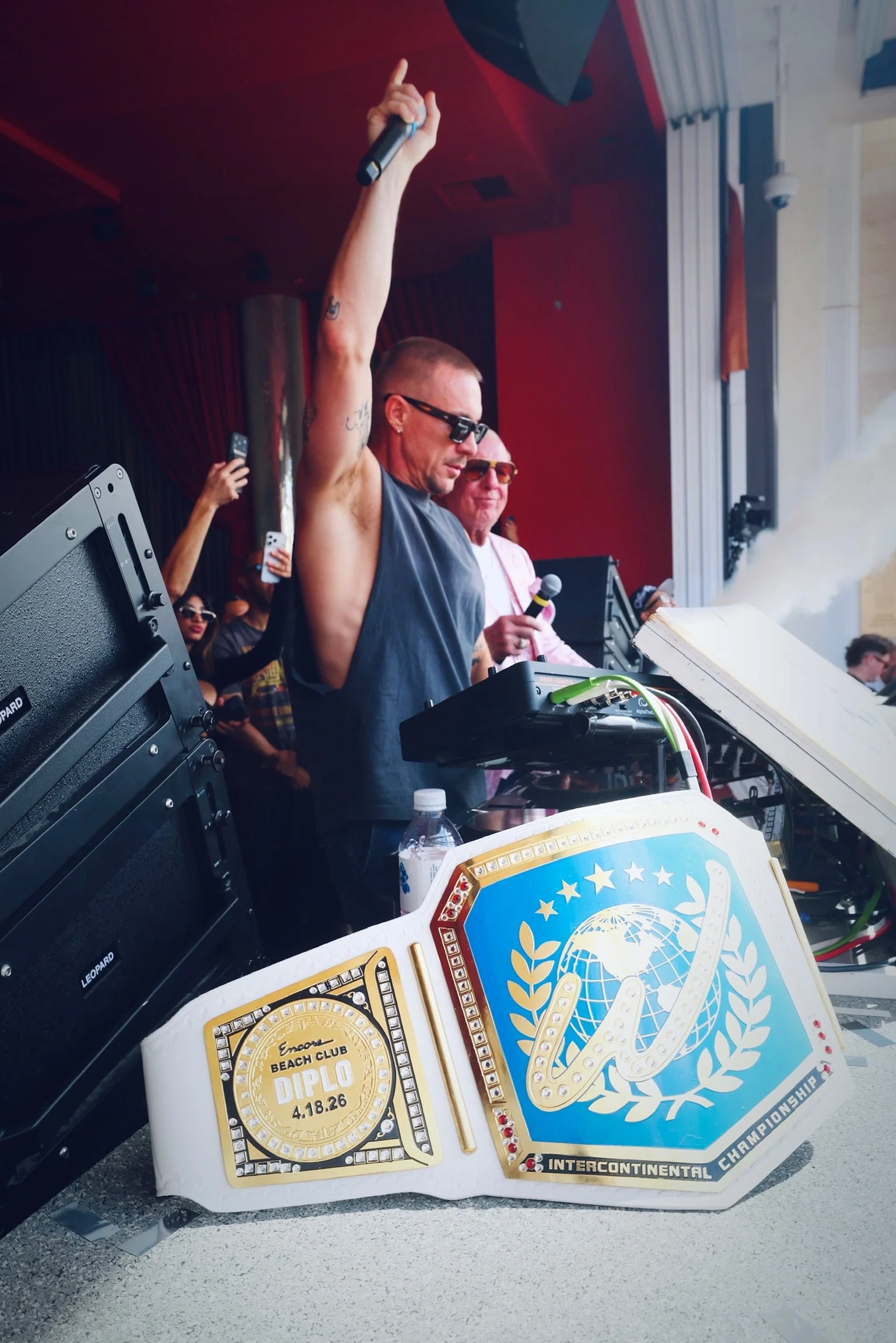  DJ Diplo stands at the Pioneer DJ decks, wearing a grey sleeveless shirt and sunglasses, looking out toward the crowd with a focused expression. In the background, Ric Flair, dressed in his signature pink patterned blazer and sunglasses, smiles broa