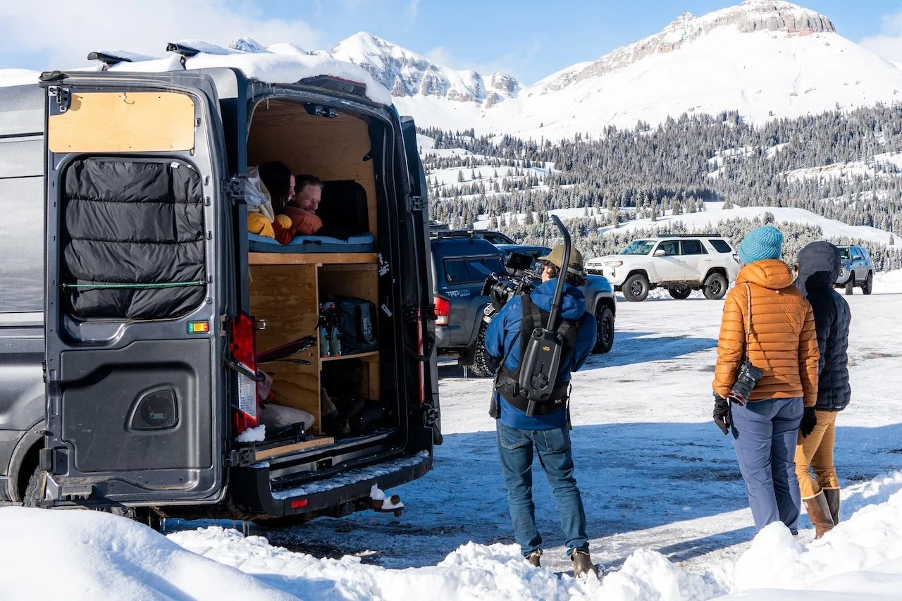 Group of people dressed in winter clothing standing outside near a camper van with an interior setup, in a snowy mountainous landscape with parked vehicles in the background.