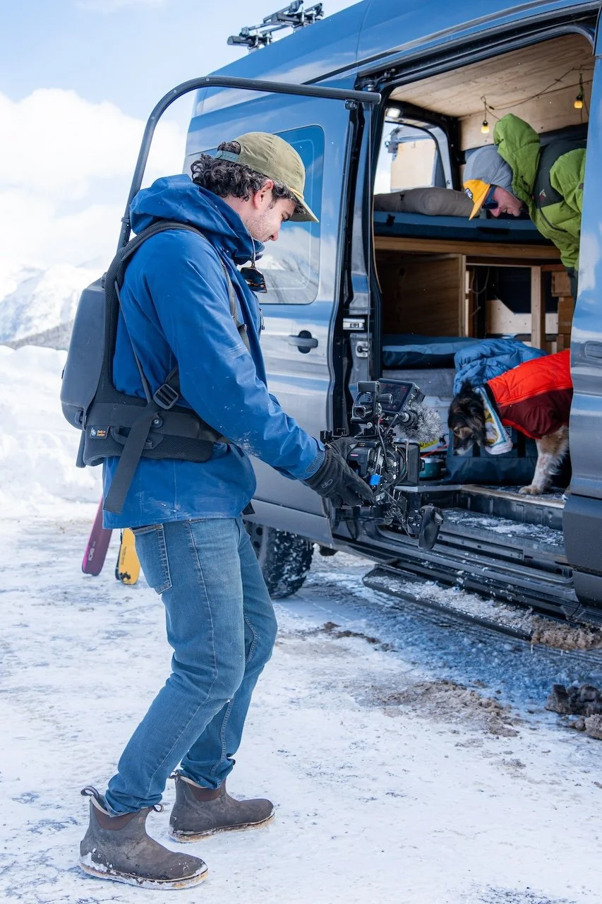 A person filming with a camera in snowy terrain near a van with an interior bed, and another person inside the van in outdoor winter clothing.