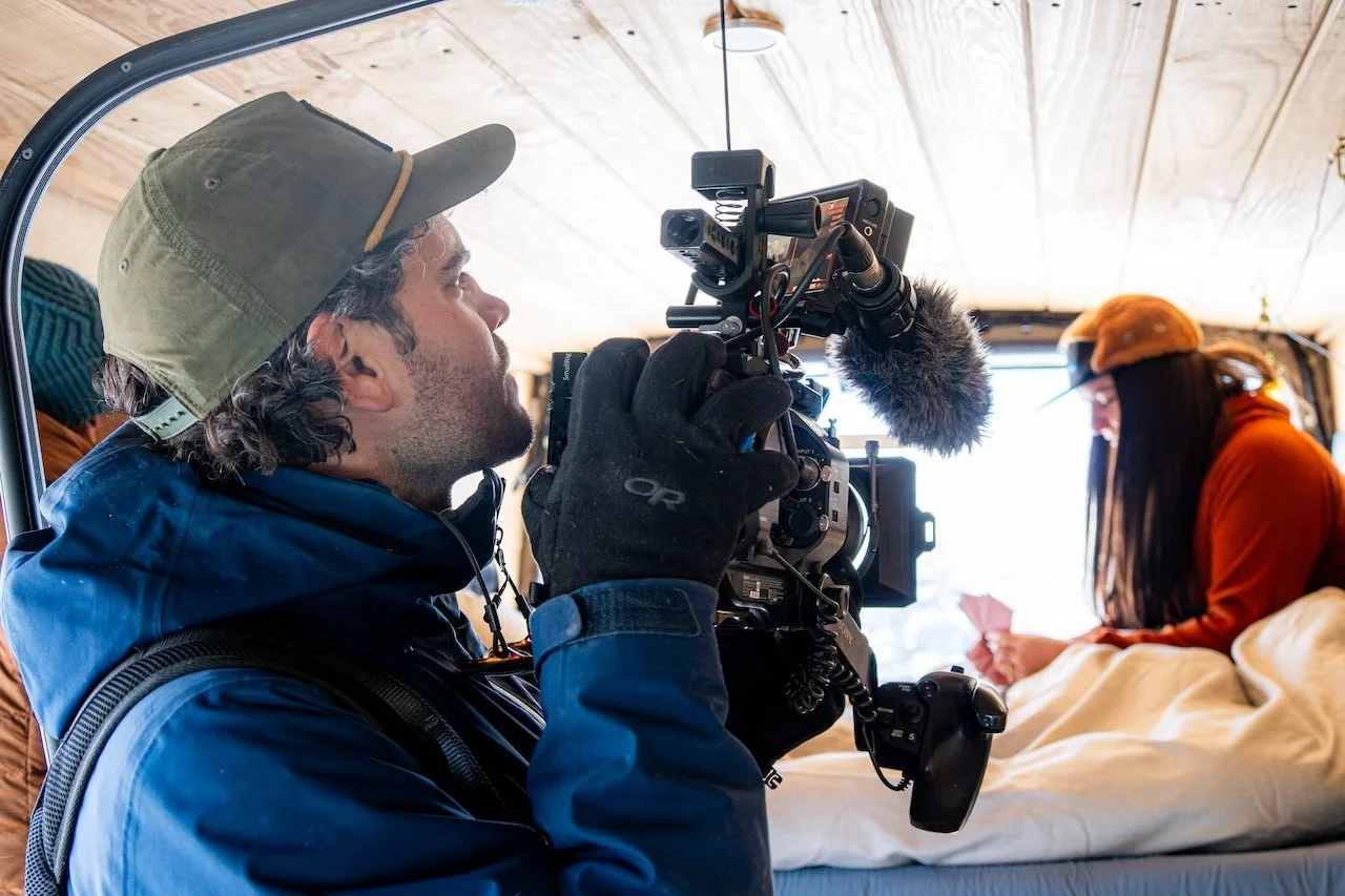 A person operating a professional video camera inside a tent, recording a woman sitting on a bed, holding a phone and smiling.